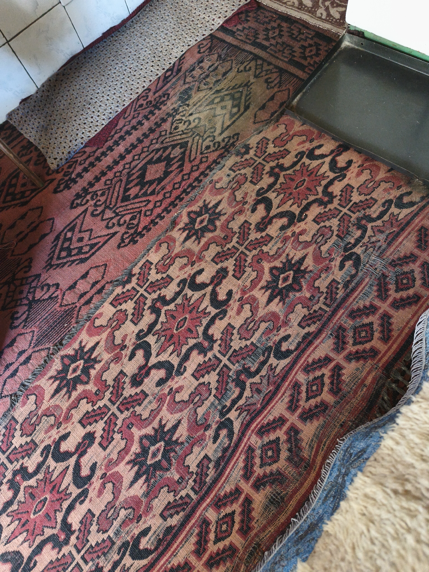The image shows a close-up of several rugs and a tiled floor. The rugs have intricate patterns in red, black, and tan. One rug is partially rolled and appears to be a textured, grey runner placed on top of a patterned rug. Another rug has a dark, rectangular object partially covering it. The floor is made of light-colored tiles. The scene appears to be an interior domestic setting, possibly a home or a shop. No people are visible in the image. The location of Slatina, Bulgaria is not evident from the image itself and cannot be inferred from the visual information provided.