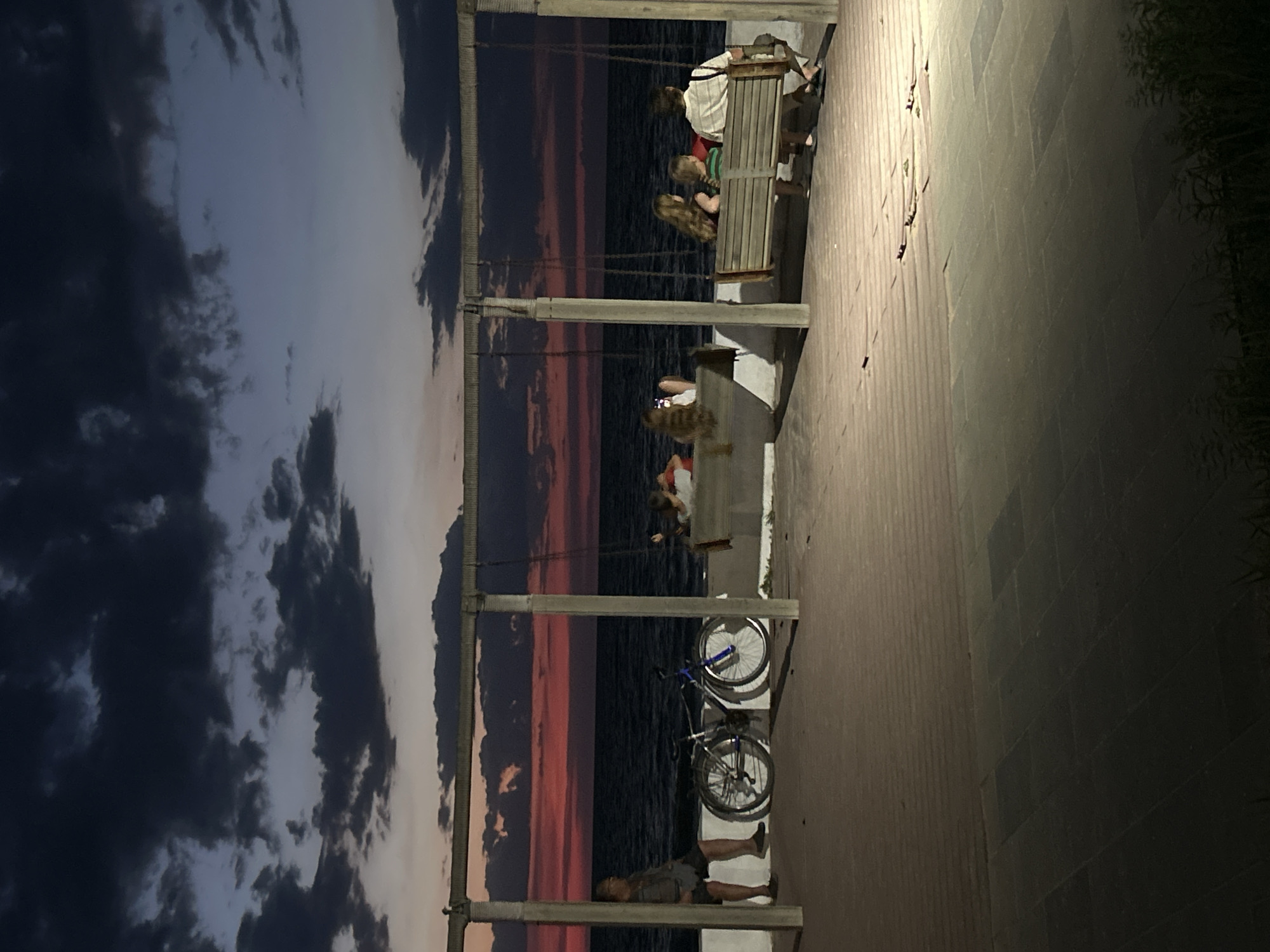 The image depicts a twilight scene at a seaside promenade in Shchelkino, Ukraine. A man is seated on a low wall to the left, with a bicycle leaning against the wall beside him. In the background, the sea is visible under a dramatic sky filled with dark clouds and a band of vibrant red and orange sunset colors on the horizon. Several people are seated on swings. On the left swing, a person is holding a smartphone. On the right swing, a family of four is seated, facing away from the camera, towards the sea. The promenade is paved with light-colored tiles, with some darker tiled areas. Artificial lighting casts illuminated patches on the ground. The overall atmosphere is calm and serene, with the fading light of day creating a picturesque view.