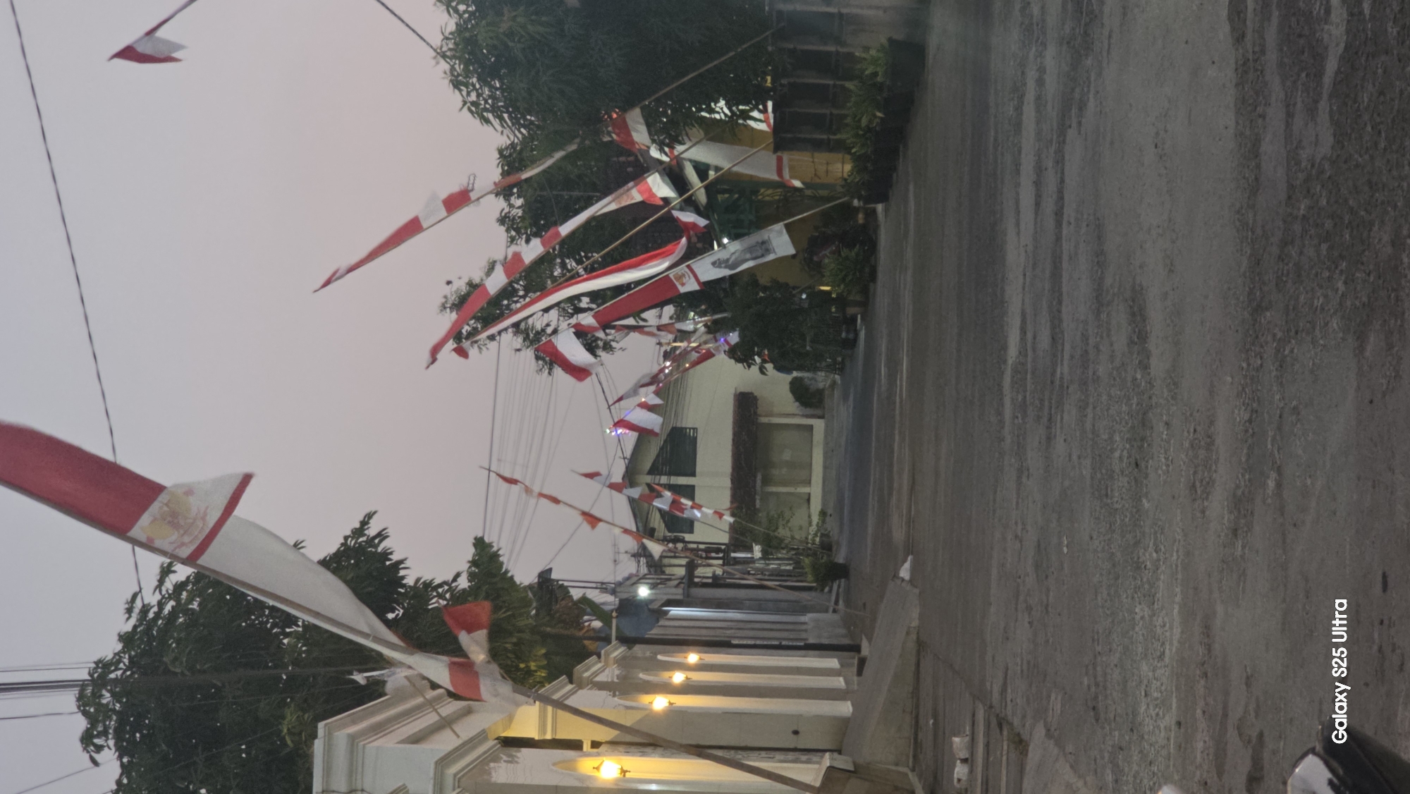 The image depicts a street in Juwana, Indonesia, lined with red and white flags, likely celebrating Indonesian Independence Day. The flags are displayed on poles along both sides of the road, with some draped across wires overhead. The street is paved and appears to be a residential or commercial area. On the left, a white building with ornate columns and lighting is visible. Further down the street, houses and trees can be seen. The sky is overcast and the lighting suggests it is either early morning or late evening. There are no people visible in the image.