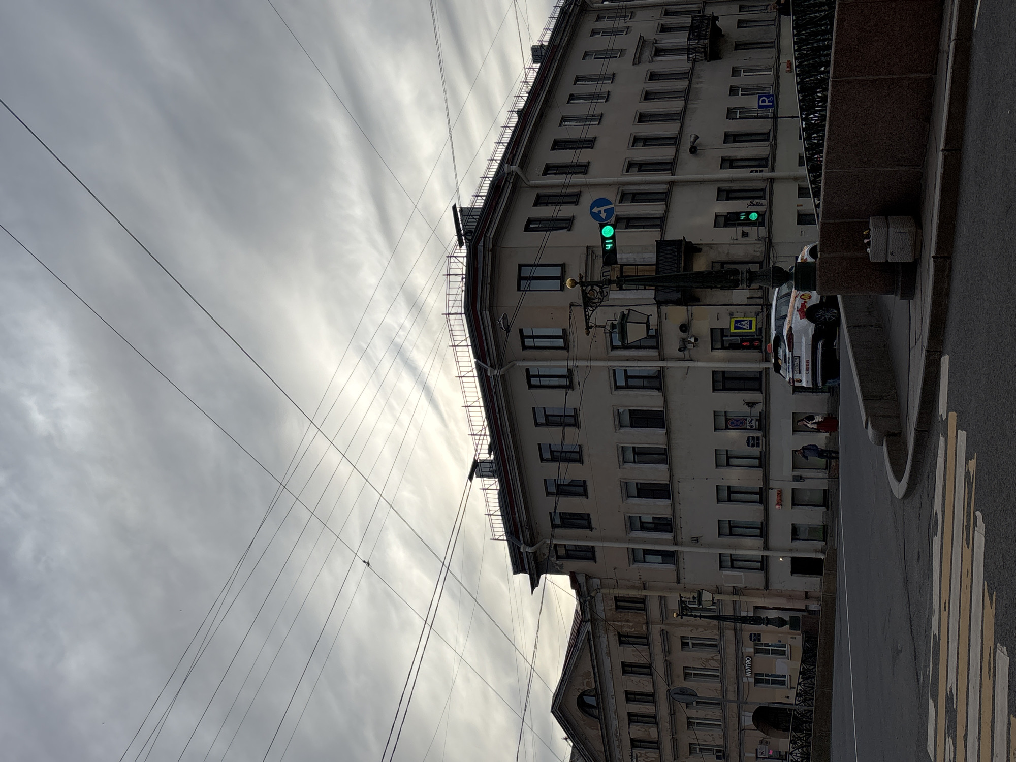 The image captures an urban street scene in Saint Petersburg, Russia, under an overcast, cloudy sky. The perspective is looking along a street, with tall, multi-story buildings lining the right side. These buildings feature numerous uniformly spaced, dark-framed windows, and one building has a distinctive rounded corner. Scaffolding is visible along the top edge of one structure.
Prominent in the frame are numerous overhead power or trolleybus lines that crisscross the sky, creating a dense network above the street. A bright green traffic light is active, positioned on a pole in front of one of the buildings. Nearby, a blue circular road sign with a white arrow indicates a left turn. Further down the street, a blue square "P" sign for parking is visible on a building facade.
The foreground on the right side includes a dark grey paved road with a white and yellow striped zebra crossing, indicating a pedestrian walkway. A white car, identified as a Yandex Taxi by the "Яндекс" and other Cyrillic text on its side, is visible near the crossing. A yellow triangular sign for a pedestrian crossing is also present. A dark, ornate railing and a sturdy stone barrier separate the street from the buildings on the right.
Visible text on building signs includes "INVITRO" (a medical laboratory) and "МИНИМУМ" (likely indicating a store or service). A few blurry figures, presumably pedestrians, can be discerned near building entrances in the background. The scene depicts a typical daytime street with active urban infrastructure, suggesting normal city life under a dull, cloudy sky.