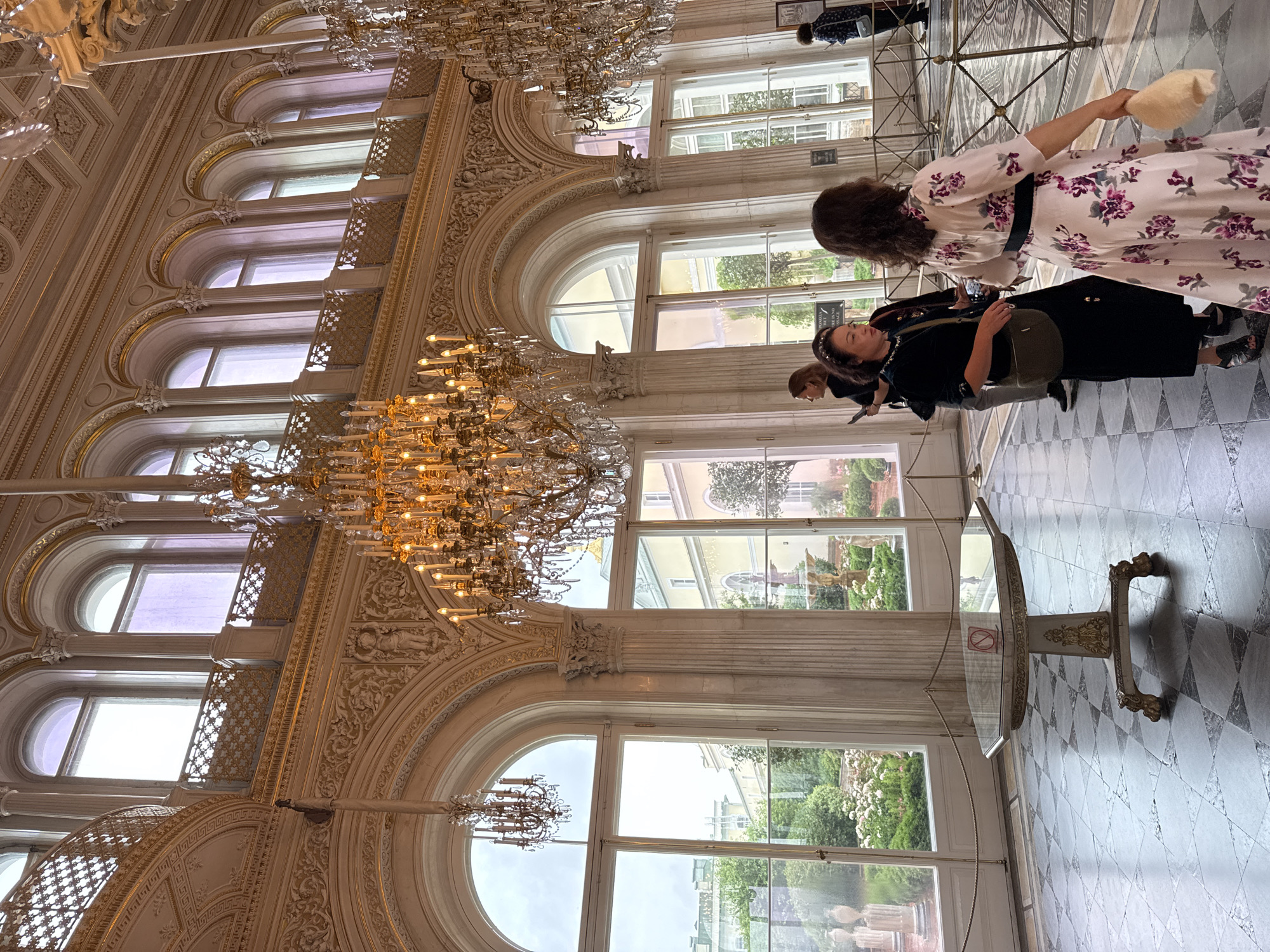 The image depicts an opulent interior with people in what appears to be a grand hall or museum. The scene is characterized by tall, arched windows that offer views of an outdoor garden or courtyard with greenery and blooming flowers. Ornate chandeliers hang from the high ceilings, illuminating the space with a warm glow. The floor is a checkered pattern of light and dark marble tiles.
Several people are visible, mostly from behind or in profile, dressed in casual attire. One woman in the foreground wears a light-colored dress with a floral pattern of pink and purple flowers, cinched with a dark belt. She is holding a light-colored, fluffy object in her hand. Another woman next to her wears a dark velvet outfit and carries a handbag. A third person, partially visible, is in the background. They appear to be casually observing the surroundings or moving through the hall.
The architectural style is classical, with elaborate moldings, columns, and decorative carvings adorning the walls and window frames. The overall impression is one of elegance and historical grandeur. The time of day seems to be daytime, with bright light filtering through the windows, suggesting either a sunny or overcast day. The location cues, such as the architecture and the presence of a formal garden, combined with the provided location context of Saint Petersburg, Russia, suggest a setting like the Hermitage Museum or another historic palace in the city. No specific emotions are overtly conveyed, but the people appear to be calmly navigating the space, likely as visitors. There is a small, circular sign with a red diagonal line and a cigarette symbol on a rope barrier near a glass table, indicating no smoking.