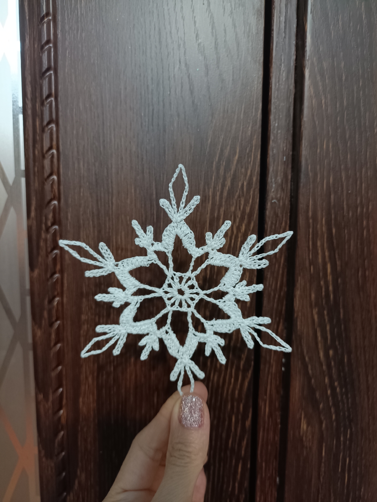 A white crocheted snowflake ornament is held by a person's hand against a dark wooden background. The snowflake has six points, each with intricate lacy details. The person's fingernails are painted with a sparkly pink glitter polish. The wooden background appears to be a piece of furniture, possibly a cabinet door, with vertical paneling and a wood grain texture. The environment is indoors. No information about the city of Bataysk, Russia, is present in the image.