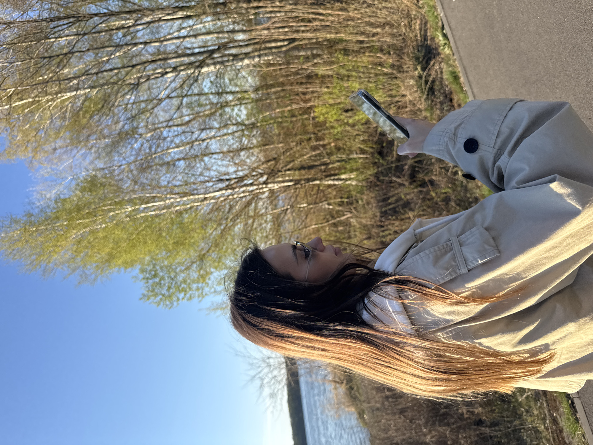 A young woman with long brown hair is standing outdoors, holding a smartphone in her left hand and looking at it. She is wearing glasses and a light beige jacket over a white shirt. The background features trees with light green leaves against a blue sky. A body of water is visible in the lower left corner. The image appears to be taken on a sunny day, possibly in a park or nature area. The location is in the city of Białobrzegi, Poland.