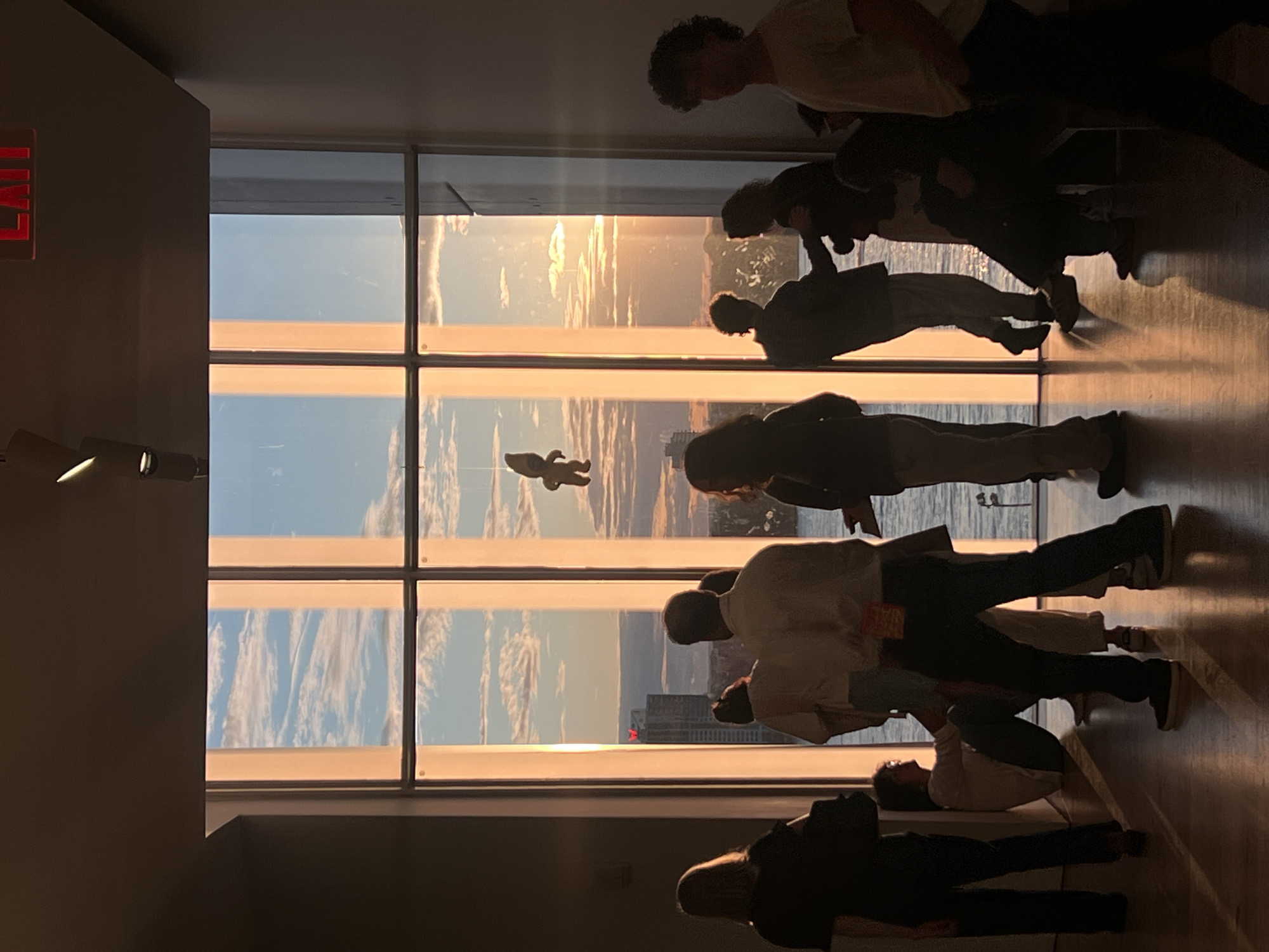 The photo shows a group of people inside a building, possibly an observation deck, looking out of a large window. The window offers a panoramic view of a city skyline during sunset. The sky is a mix of blues, oranges, and yellows. The cityscape includes tall buildings and a glimpse of water. There is an "EXIT" sign on the left side of the wall and a few track lights. A child's toy is seen stuck to the window. Given the tall buildings and sunset view, the city is likely New York City, USA. The people are standing near the window and seem to be observing the sunset. They appear to be in a public place with many people.