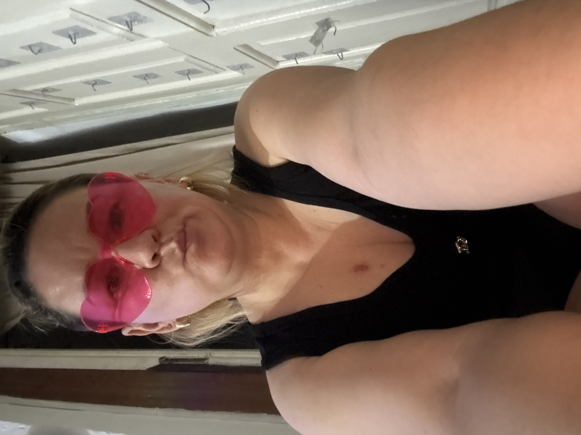 In Chardara, Uzbekistan, a woman is photographed in a self-portrait. She is wearing a black top and pink heart-shaped glasses. Her hair is blonde and pulled back. The background appears to be indoors, with a white door and some decorative elements. The lighting suggests it is evening or an indoor setting.