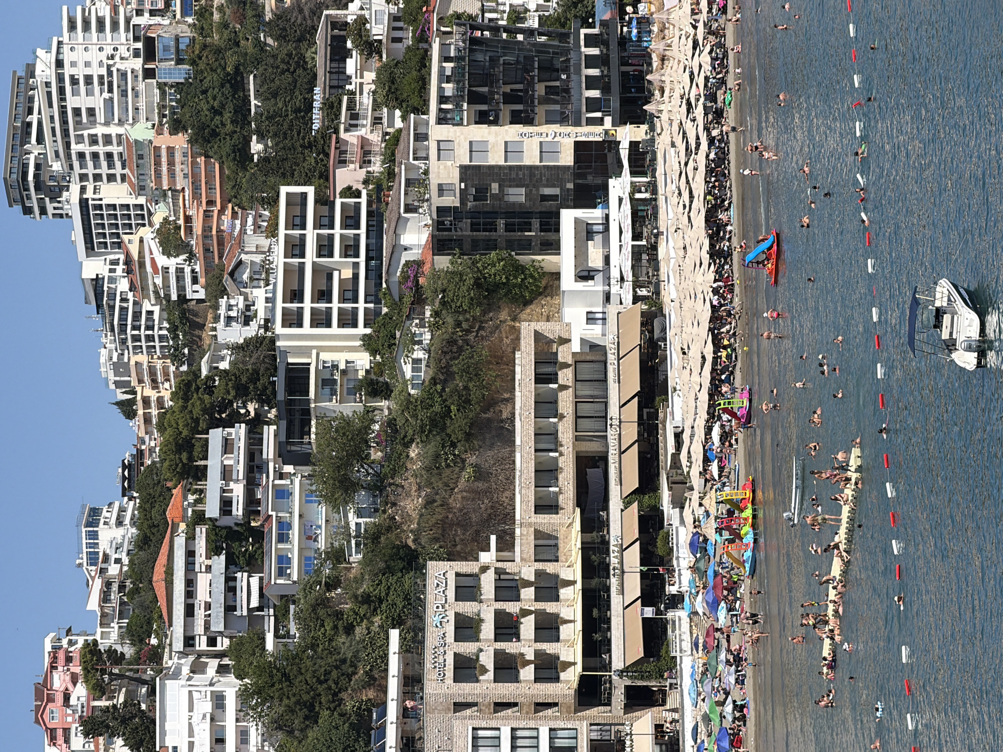 The image depicts a bustling beach scene in Ulcinj, Montenegro, with a densely populated hillside covered in hotels and residential buildings overlooking the coastline. The foreground is dominated by the turquoise waters of the Adriatic Sea, where numerous people are swimming, lounging on floating platforms, and enjoying water activities. A small white motorboat is docked in the lower right portion of the frame.
The beach itself is a vibrant spectacle, packed with umbrellas of various colors providing shade for sunbathers. Several colorful inflatable water slides and toys are present, indicating a family-friendly atmosphere. People are seen walking along the shoreline, relaxing on lounge chairs, and engaging in beach activities.
Behind the beach, a strip of buildings includes restaurants and cafes with outdoor seating, sheltered by beige awnings. A prominent stone-clad hotel, "HOTEL & SPA PLAZA," with multiple floors and balconies, stands out.
The background reveals a steep hillside densely packed with a multitude of modern and older buildings, a characteristic of Ulcinj's architecture. These structures, ranging from apartment complexes to hotels, cascade down the slope towards the sea, creating a dramatic urban landscape against the clear blue sky. Green vegetation interspersed among the buildings suggests a Mediterranean climate. The overall impression is one of a lively tourist destination during peak season.