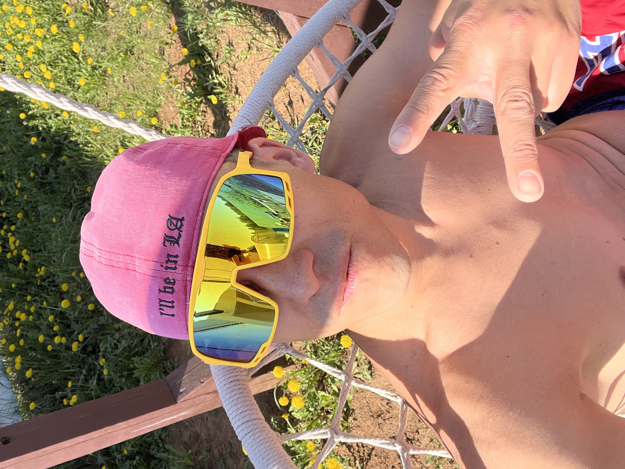 A man is reclining in a rope hammock outdoors on a sunny day. He is wearing a pink baseball cap with black lettering, yellow-framed sunglasses, and red and blue patterned shorts. He is shirtless. His right hand is raised, fingers extended, making the "peace" sign. A field of yellow flowers is visible in the background. The location is Chaikovsky, Russia.