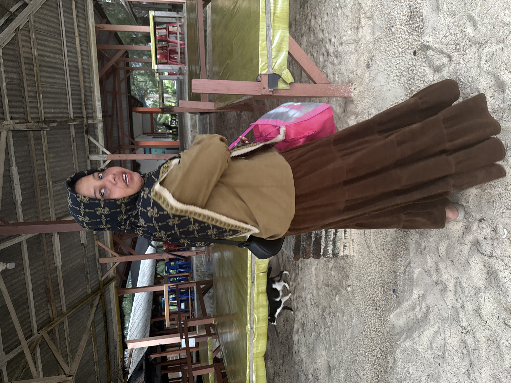 A full-shot, eye-level, outdoor medium-light natural light portrait shows a young Indonesian woman, wearing a black and gold patterned hijab and a brown tiered skirt, standing on a sandy surface under a covered wooden structure. She faces the camera with her head turned to her right, her mouth slightly agape. A brown jacket is worn over a light-colored garment, and a black shoulder bag is slung across her right shoulder. A bright pink tote bag hangs from her right hand. Behind her, a black and white cat walks away from the camera towards the left. The background reveals a sandy area with scattered debris, a brick pillar on the right, and a raised wooden platform with yellow coverings. Further back, a wooden structure with a corrugated roof and exposed beams is visible, as well as rows of red chairs and tables. The location is identified as Meunasahbale, Indonesia.