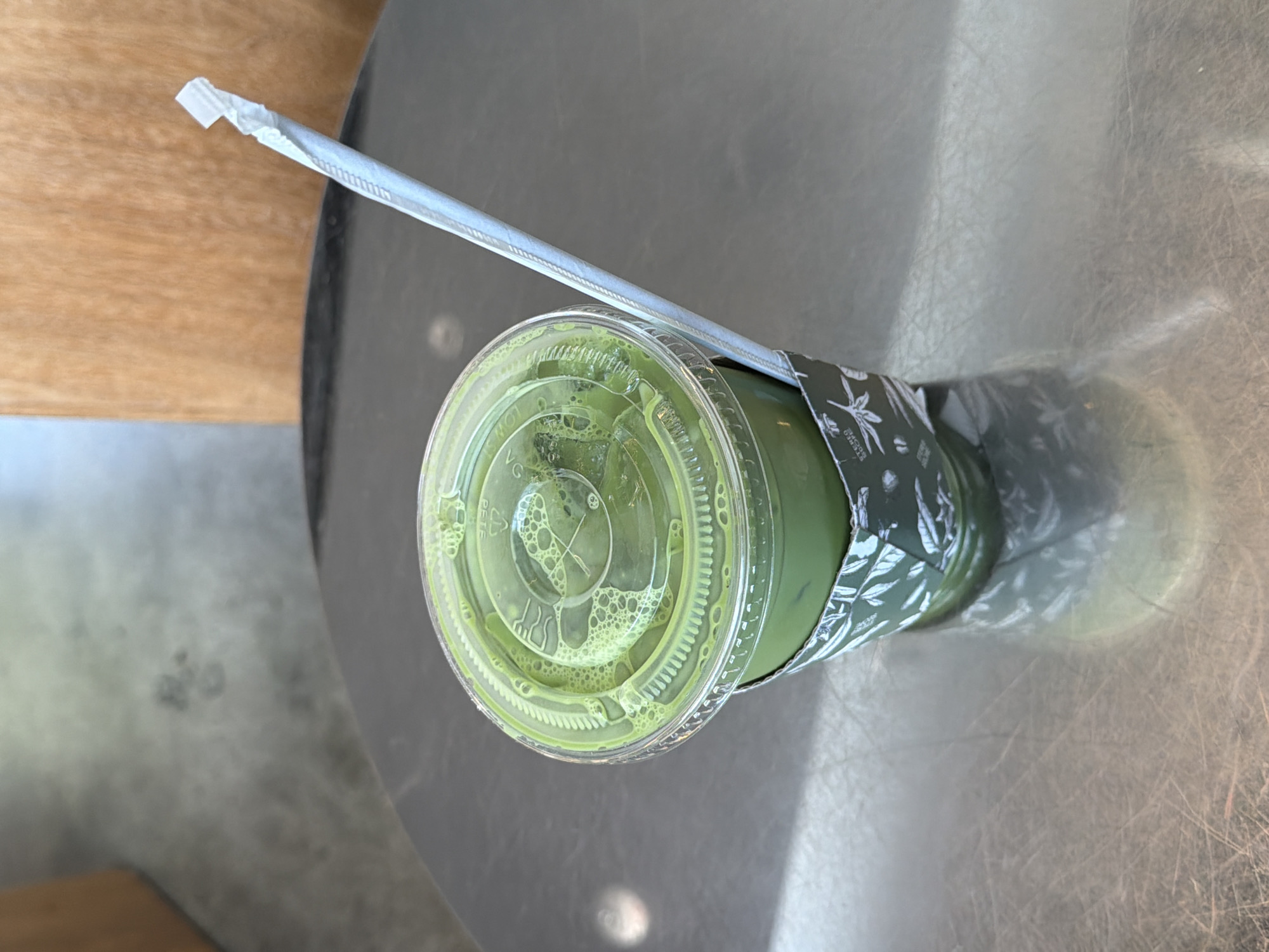 The image shows an overhead shot of a green-colored drink in a clear plastic cup with a clear plastic lid. A white paper straw sits in the cup through a hole in the lid. The cup has a black and white sleeve with a leaf pattern. The cup sits on a gray surface. The environment appears to be indoors, possibly a cafe or restaurant. Based on the visible signs, it is impossible to pinpoint an exact location within Los Angeles. The presence of the drink suggests someone is, or was, present to consume it.