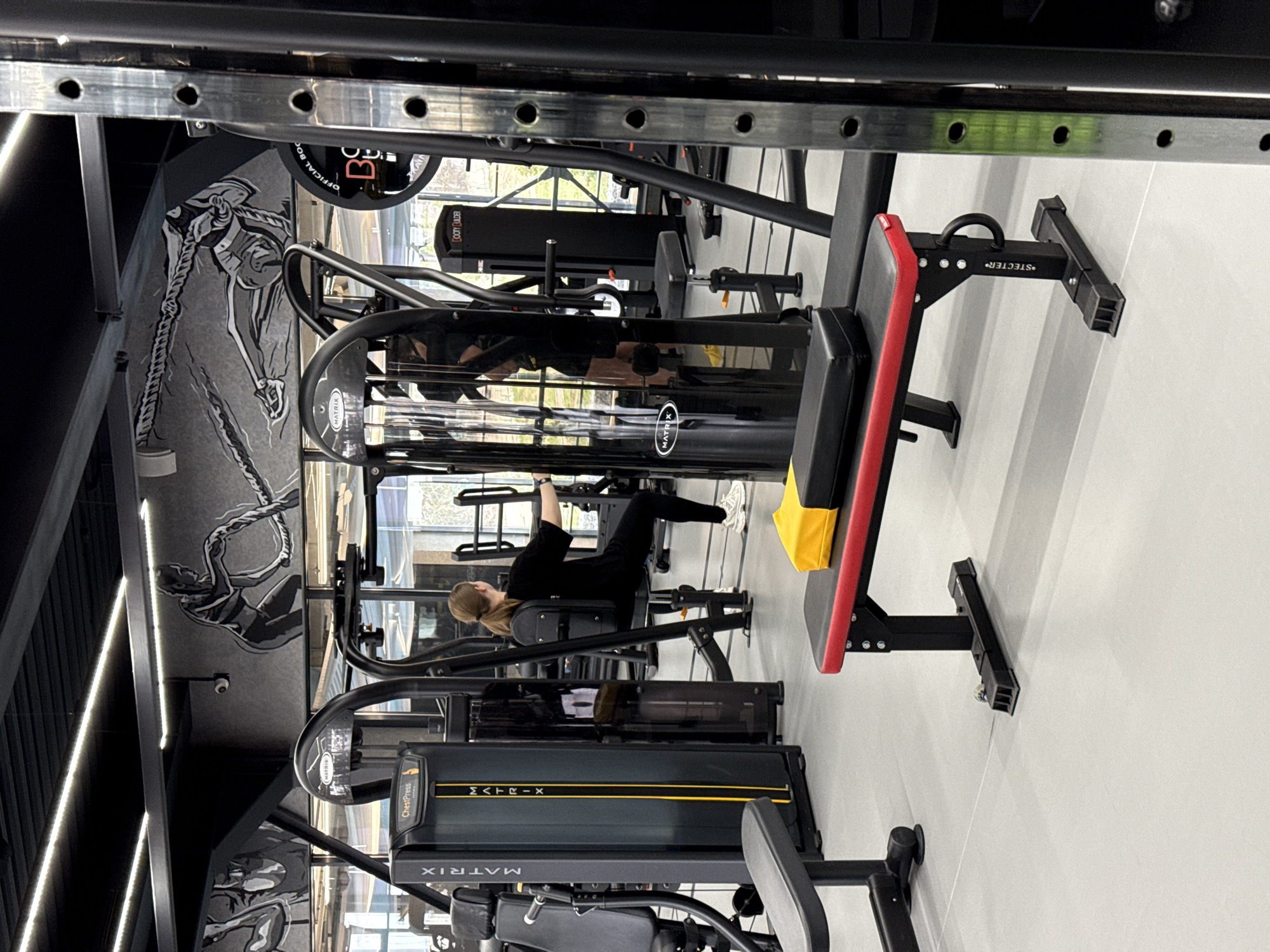 The image, rotated 90 degrees clockwise, depicts the interior of a modern gym or fitness center in Votkinsk, Russia. The setting is brightly lit with artificial strip lighting and features light-colored walls adorned with striking black and white murals of athletes, including one illustrating a person climbing a rope.
A single individual, a person with light brown hair tied back, is visible in the mid-ground, actively engaged in a workout. They are wearing all-black athletic attire and white sneakers, using a black gym machine for what appears to be a strength training exercise, performed with focus and determination.
The facility is equipped with a variety of contemporary exercise machines. Several pieces of equipment are clearly branded "MATRIX," with one specifically labeled "ChestPress." Other visible items include a red and black weight bench with a yellow towel draped over it, a machine identified as a "BOOTY BUILDER," and a black attachment bearing the name "STECTER." A large metal rack system is prominent in the foreground. Partial text on a weight plate reads "OFFICIAL BOO" and the number "30." Large windows in the background offer glimpses of the outside, contributing natural light to the indoor environment. A small security camera is also mounted on a wall.
