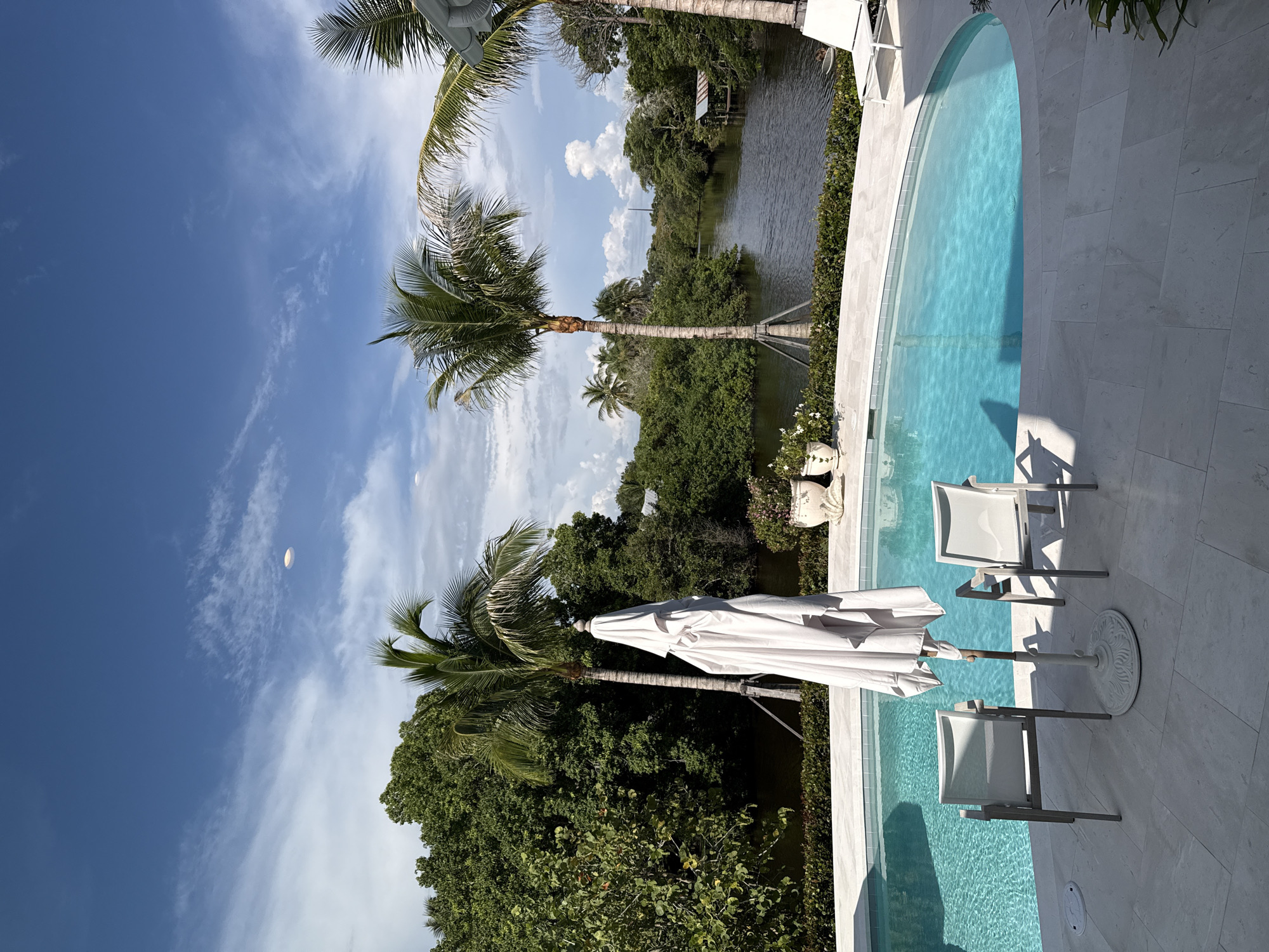 This image depicts a backyard swimming pool area in East Naples, Florida, United States of America. The scene includes an oval-shaped swimming pool filled with clear blue water, surrounded by a light-colored paved patio. Two white chairs with mesh seats and a closed white umbrella on a stand are positioned on the patio near the pool. In the background, lush green trees and palm trees line a dark body of water, likely a canal or estuary. A small rustic structure with a roof is visible on the opposite bank of the water. The sky is a bright blue with scattered white clouds. There are no people visible in the image.