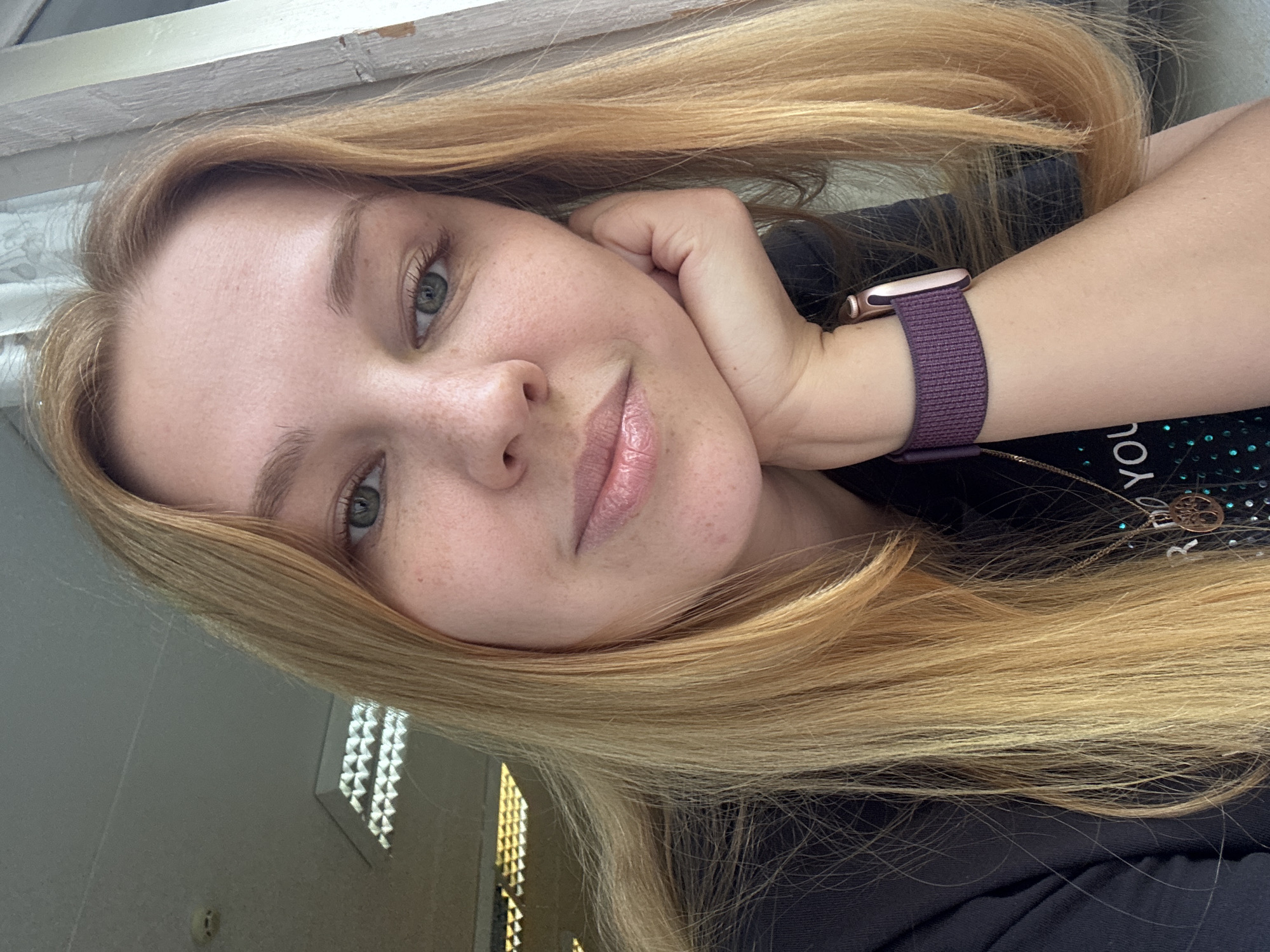 A fair-skinned woman with blonde hair and freckles is posing for a selfie. She is resting her head on her hand, and wearing a purple watch. She is wearing a black t-shirt. The background includes a window with white frames and what appears to be an office environment with lighting fixtures. The location appears to be indoors. No specific details indicate Rusanovo, Russia.