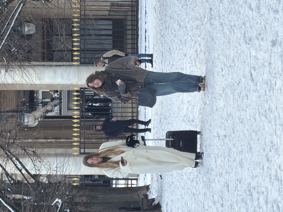 This image depicts a snow-covered scene within the Palais Royal gardens in Paris, France. The ground is uniformly covered in white snow, indicating a winter setting. In the foreground, two women walk from left to right across the snow.
The woman on the left has long, light brown hair and is wearing a long, double-breasted white coat, dark pants, and dark shoes. She holds a white smartphone in her left hand, looking down at it, and has a small black handbag with a prominent "CC" logo on her right shoulder. She is pulling a black rolling suitcase with an extended silver handle.
The woman on the right has shoulder-length brown, wavy hair and is dressed in a plaid jacket, a dark scarf, blue jeans, and light-colored sneakers. She holds a white smartphone in her right hand and carries a dark blue or black tote bag on her right shoulder. She is looking slightly upward and to her right.
In the background, behind a black wrought-iron fence adorned with golden spear-top finials, stands a grand building with prominent classical columns and recessed windows. Several streetlights are visible hanging from the building's facade. Behind the fence, two additional men are present on a paved area. A man in a dark hooded sweatshirt and dark pants stands centrally, with his arms slightly out. To his right, another man with grey hair, wearing a light-colored vest over a dark long-sleeved shirt and blue jeans, stands looking towards the two women in the foreground. The bare branches of trees are visible on the left side of the frame.