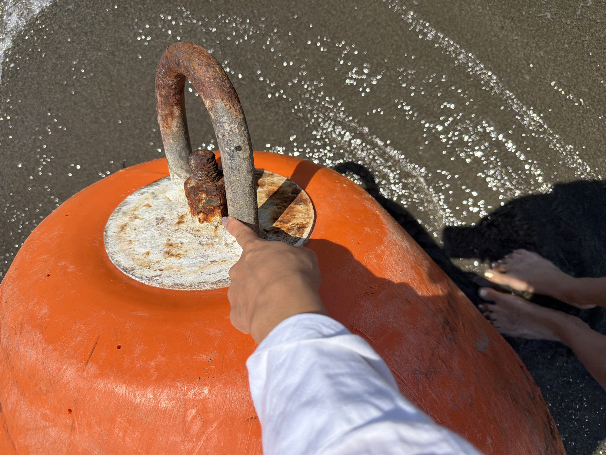 A person's left hand with white long-sleeved shirt is seen pointing at a rusty metal loop attached to an orange buoy, which is partially submerged in dark wet sand. The sandy area is scattered with shimmering reflections of light, possibly from water. In the lower left of the frame, the person's bare feet are visible standing in the sand. The scene appears to be on a beach near Meneou, Cyprus.