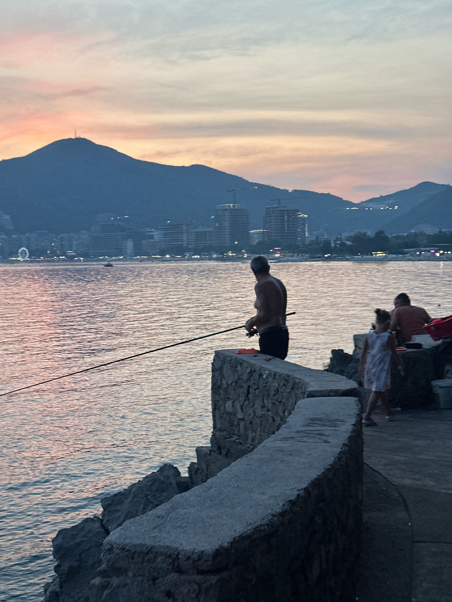 A serene sunset scene unfolds in Budva, Montenegro, featuring a calm sea with gentle ripples reflecting the pastel hues of the sky. In the foreground, a man stands on a stone embankment, actively fishing with a rod. To his right, a young girl in a patterned dress walks away from the camera, while a person in an orange shirt sits further back. Across the water, the Budva skyline is visible, with buildings silhouetted against the fading light, and a prominent mountain looms in the background, topped with a structure. The overall atmosphere is peaceful, indicative of an evening by the coast.