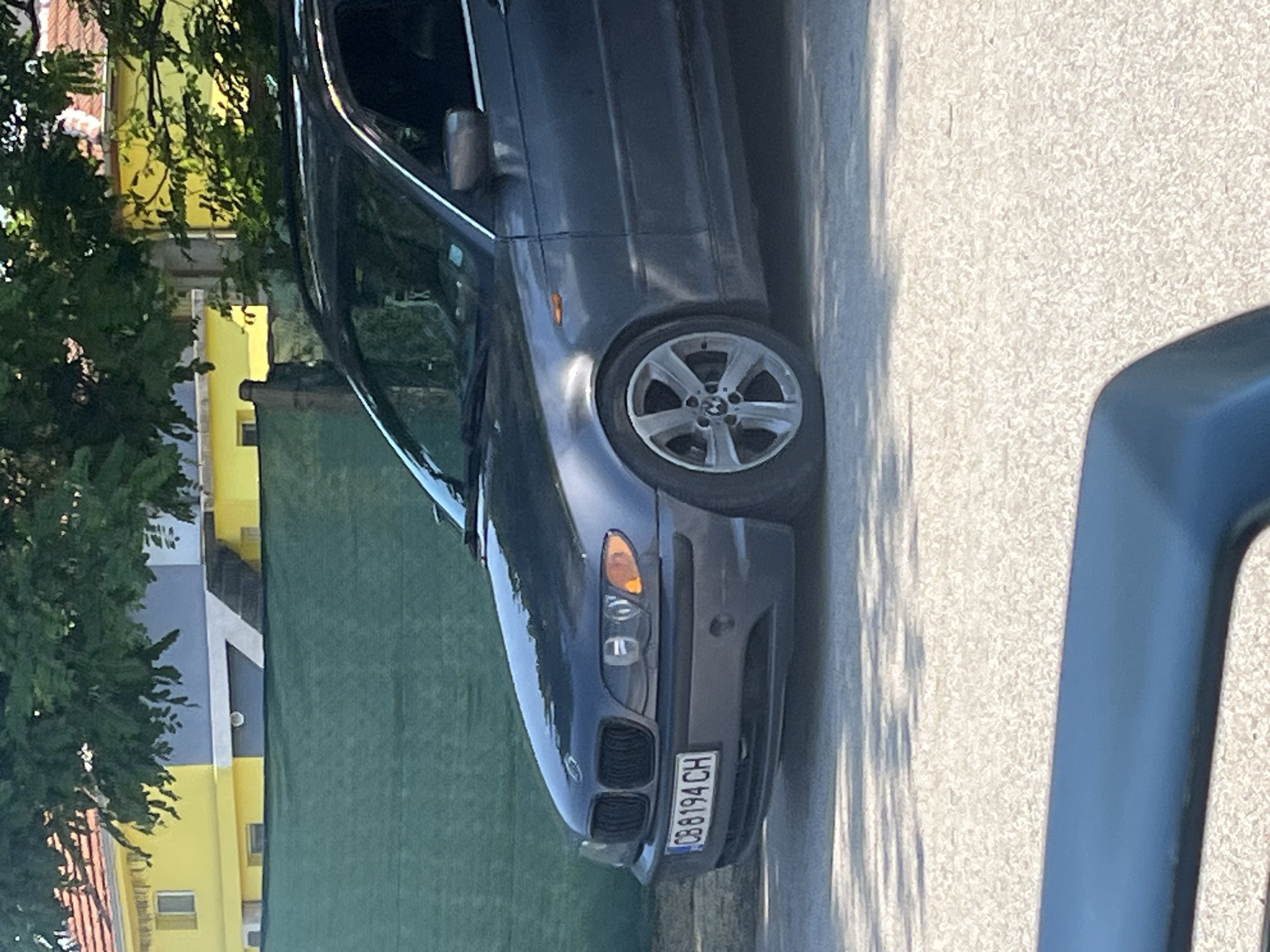 A gray BMW sedan is parked on a paved surface adjacent to a green-painted wall and a tree in Trilistnik, Bulgaria. The front of the car, with license plate "CB 8194 CH", is visible. A portion of a building painted in yellow and gray can be seen in the background behind the tree.