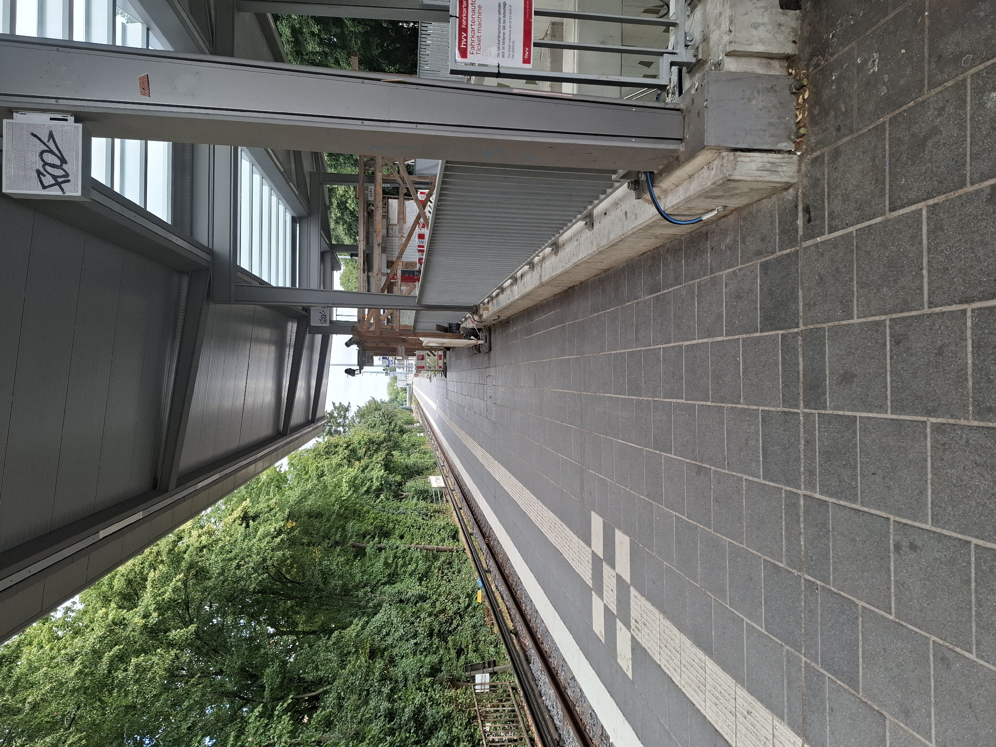 The image depicts an outdoor public transit platform, likely a train or tram station, with a covered waiting area. The platform is paved with dark gray square tiles. To the left, train tracks run alongside the platform, bordered by a metal fence and lush green trees. A white tactile paving strip is visible along the edge of the platform.
The covered waiting area is supported by metal pillars and features a grey roof with translucent sections that allow light to filter through. There is some construction or renovation happening, with scaffolding and wooden structures visible in the background. A metal railing separates the platform from the construction area.
A sign with the text "hvV Fahrkartenautomat Ticket machine" in German and English is prominently displayed on a pillar near the right edge of the image. Below this, another sign with the "hvV" logo is visible. Graffiti, stylized as "FOOL," is present on a speaker or panel mounted on the ceiling of the waiting area.
The environment appears to be a suburban or urban setting, given the presence of public transit infrastructure and mature trees. The overall impression is one of a functional, albeit partially under construction, public space. The image does not contain any identifiable people.