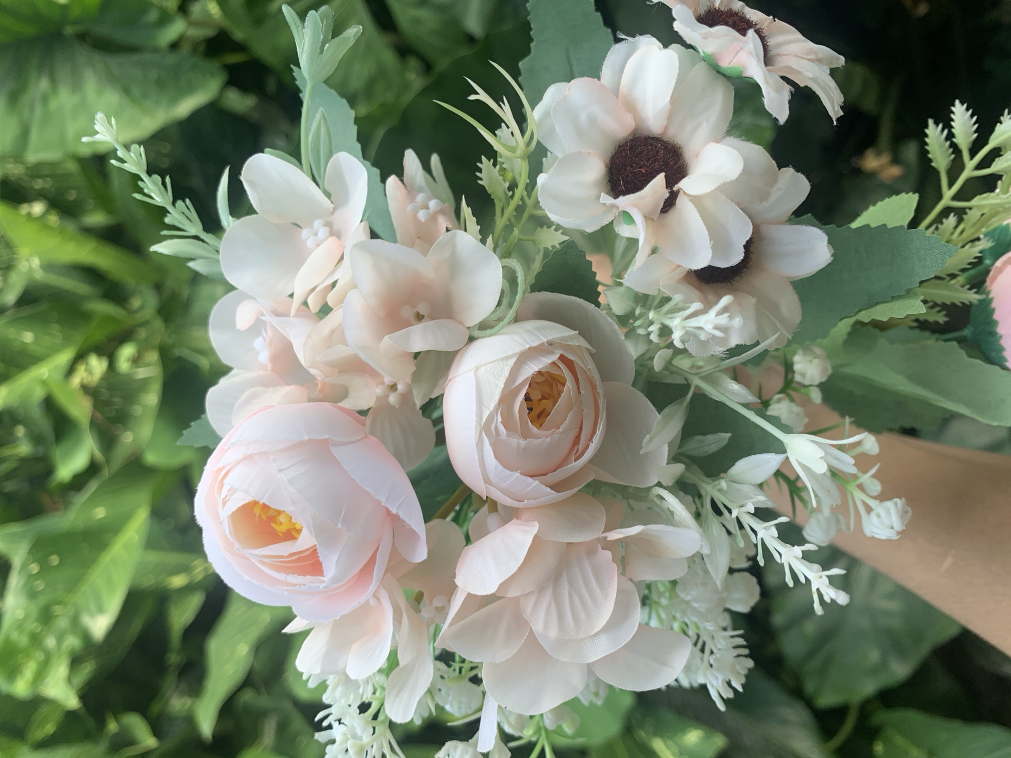 A close-up, vertical shot captures a bouquet of artificial flowers, predominantly in pale pink and white hues, held by a person's hand and forearm in the lower left of the frame. The bouquet is composed of several rose-like flowers with soft pink petals and yellow centers, interspersed with smaller white flowers resembling baby's breath and delicate, light-pink blossoms with small white centers. There are also a few daisy-like flowers with white petals and dark brown centers. Green artificial leaves and stems are visible throughout the arrangement. The background is softly blurred, revealing lush green foliage, suggesting an outdoor environment. The scene appears to be a personal, decorative arrangement, not tied to a specific public landmark or activity in Kuala Pembuang, Indonesia.