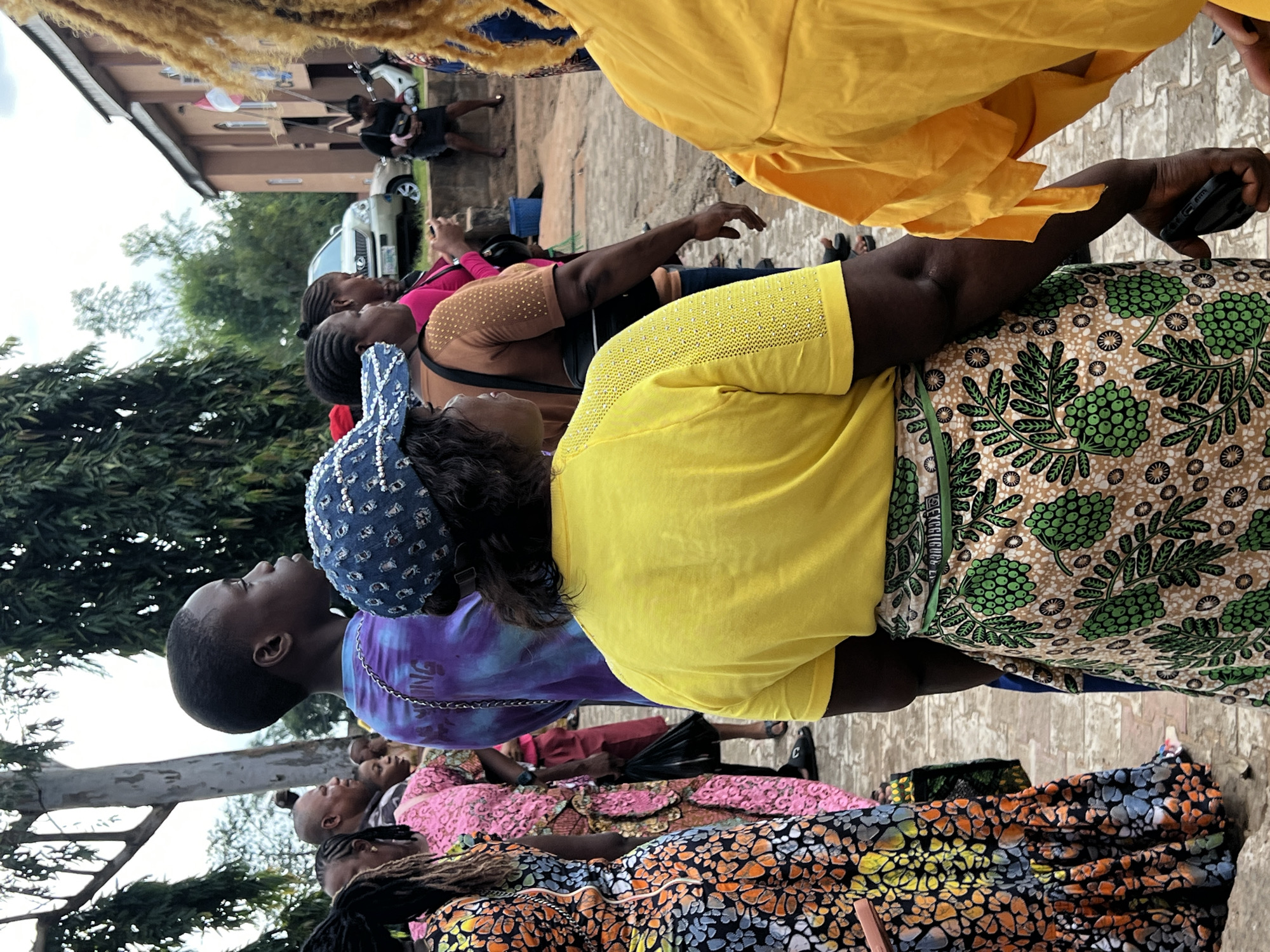 A group of people are gathered outdoors in what appears to be a street or courtyard in Nnewi, Nigeria. The central figure, a woman wearing a yellow t-shirt and a patterned skirt with green fruit motifs, is facing away from the camera. She is also wearing a blue baseball cap adorned with white embellishments. To her left, a young person in a tie-dye shirt and a person in a pink patterned dress are visible. Further to the left, another person is wearing a vibrant dress with a black, orange, and yellow pattern. In the background, a tan building with a red and white flag is visible, along with a silver SUV and a white motorcycle. The ground is paved with cobblestones or similar material. The overall atmosphere suggests a public gathering or event.