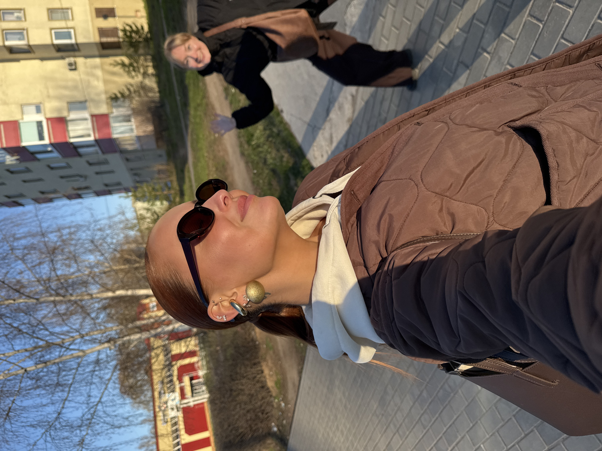 A selfie taken on a sunny day shows two women on a paved walkway in Chaikovsky, Russia. The woman in the foreground is wearing sunglasses and a brown quilted jacket over a white hoodie, with multiple earrings visible in her left ear. Behind her, another woman is walking and smiling towards the camera, wearing a black jacket and dark pants, carrying a brown bag. The background includes apartment buildings, trees, and a clear blue sky. The overall scene depicts an urban environment during daylight hours.