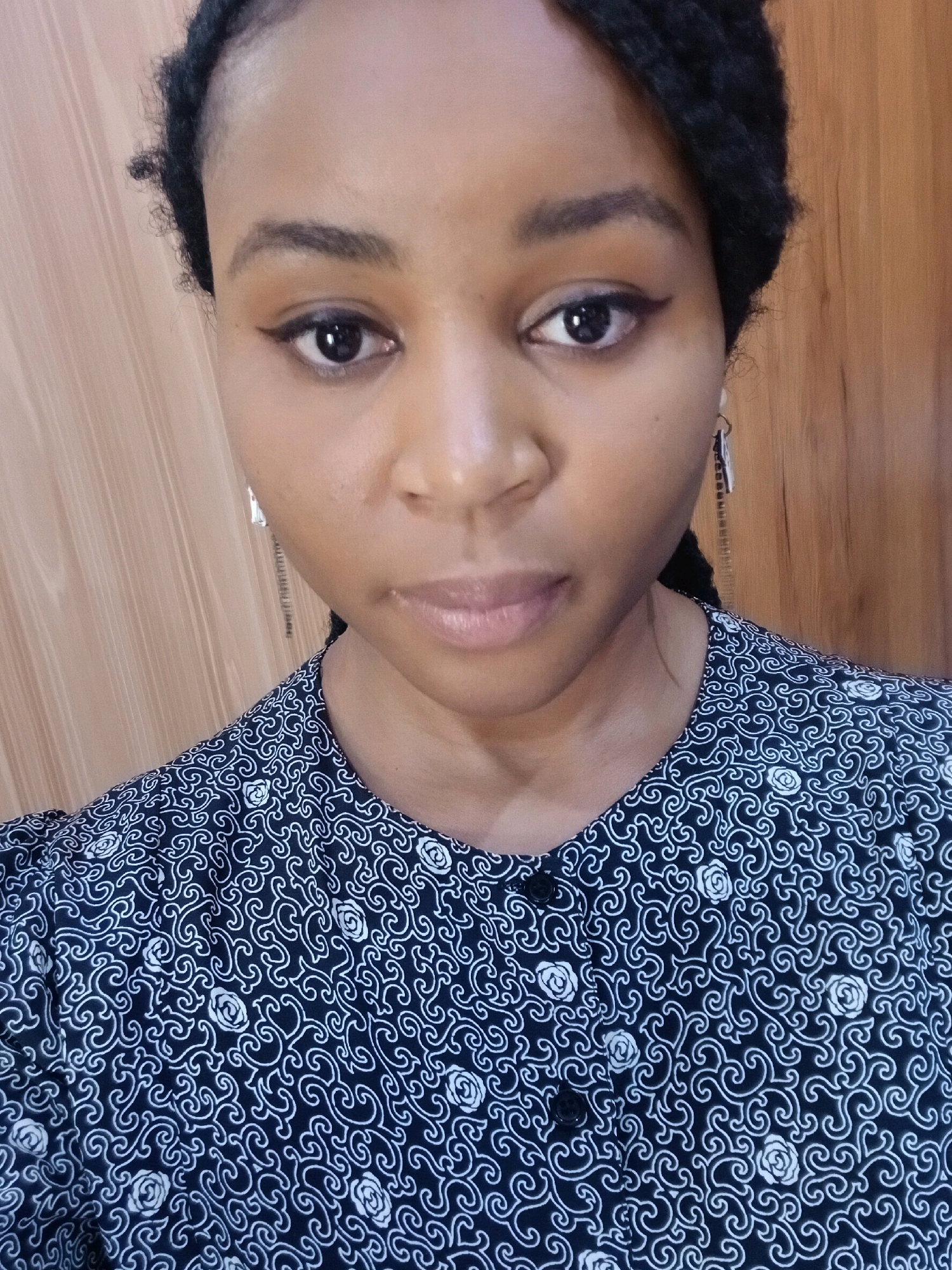 A close-up, head-and-shoulders shot captures a young Black woman with dark braided hair from the chest up. She is looking directly at the camera, her expression neutral. She wears a dark blue and white patterned blouse. Her makeup includes eyeliner and mascara. Silver earrings with a rectangular pendant are visible. The background consists of a wooden-paneled wall. The image appears to be a selfie taken indoors.