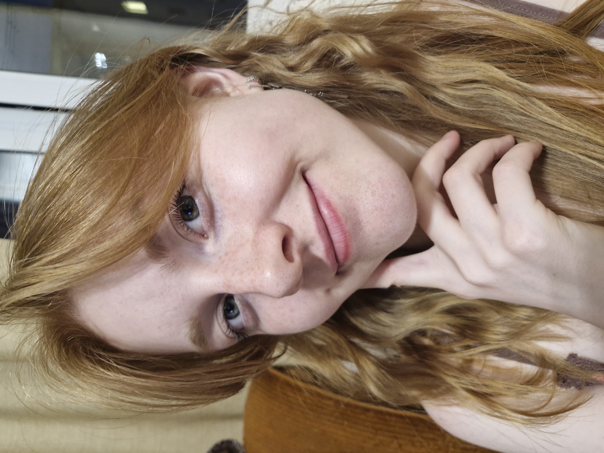 A close-up, rotated portrait features a young woman with fair skin, prominent freckles on her nose and cheeks, and long, wavy reddish-blonde hair. Her green-blue eyes are open, looking directly at the viewer, and she wears a subtle, gentle smile with her lips slightly parted. Her right hand is visible, with fingers softly resting against her face and neck, touching her hair. A small, delicate earring or ear cuff adorns her visible ear. The lighting appears natural, suggesting a daytime indoor setting. The background is softly blurred, showing indistinct interior elements and light reflections. The woman's expression conveys a sense of calm and gentle engagement. There is no visible text in the image. The location context provided is Yekaterinburg, Russia.