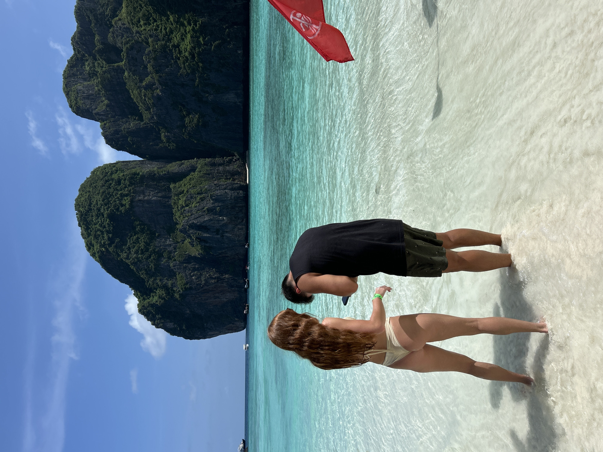 This is a medium shot, eye-level, outdoor, daytime photograph taken on a beach in Ao Nang, Thailand. In the foreground, two individuals, a woman with long, red hair wearing a beige bikini and a man with dark hair wearing a black tank top and dark shorts, stand in shallow, clear turquoise water. The woman faces away from the camera, looking towards the man, who is also facing away and looking at his phone. A red flag with a white symbol of a person with a line through it is visible in the right foreground, partially obscuring the water. In the background, the turquoise water extends to meet two large, lush green rocky islands under a clear blue sky with scattered white clouds. Several boats are visible in the distance on the water. The beach is white sand, and the water is exceptionally clear, revealing ripples and shadows on the sandy bottom.