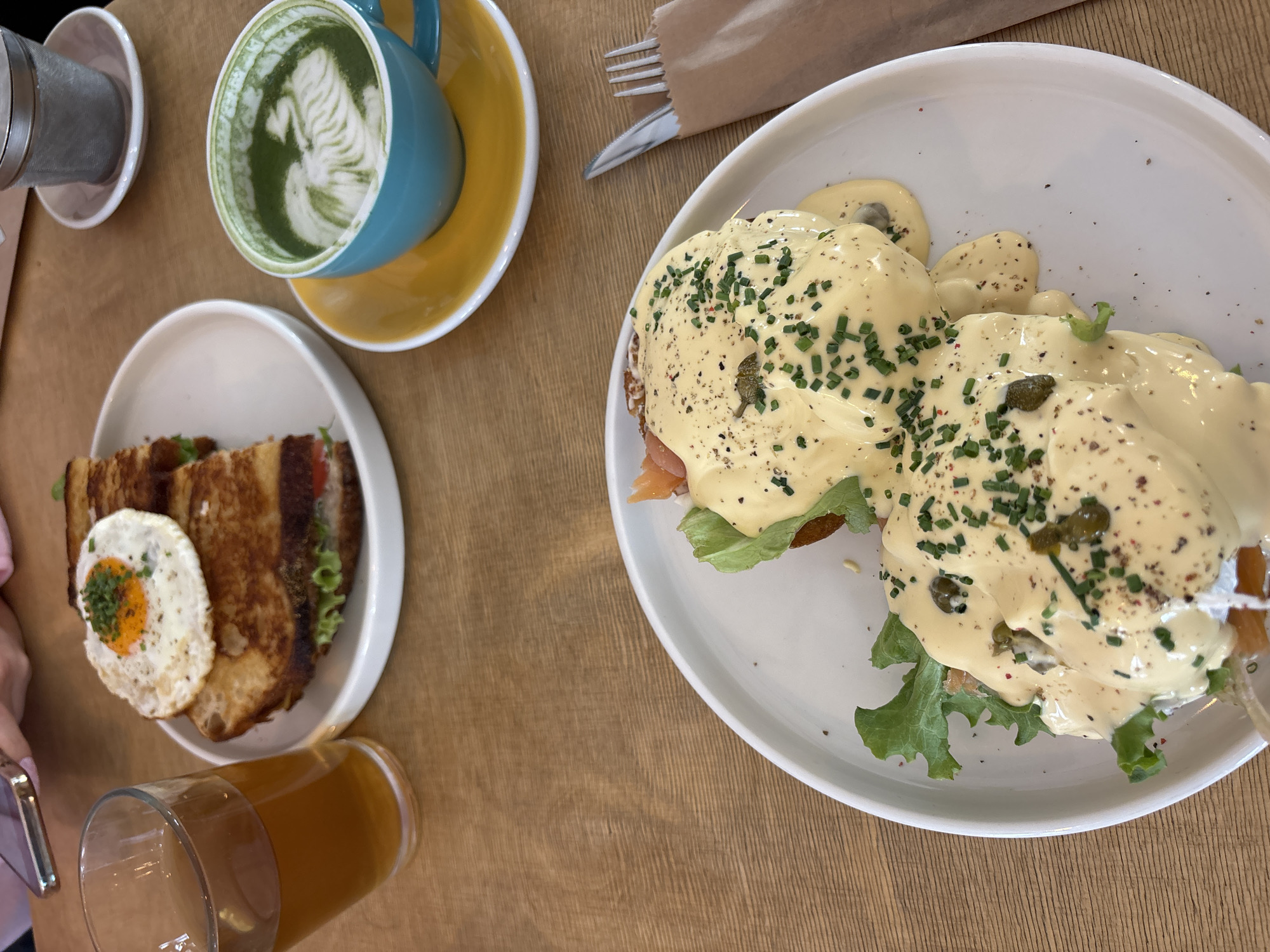 The image shows a table setting with food and drinks, likely in a cafe or restaurant. A plate of Eggs Benedict, topped with hollandaise sauce, chives, and capers, sits on the right. To the left of the Eggs Benedict is a plate with a sandwich, topped with a fried egg. A glass of amber-colored liquid, possibly beer or juice, is visible near the sandwich. Above the sandwich is a blue cup of matcha latte on a yellow saucer. A silver tea infuser sits next to the cup, and a knife and fork in a brown paper bag are to the right of the cup. The table has a light brown woodgrain pattern. No people are clearly visible, although a hand holding a cell phone is partially visible in the lower left corner. Given the food style and ambiance, it's plausible the image was taken in a cosmopolitan cafe in Varna, Bulgaria, but there is no visual confirmation in the image.