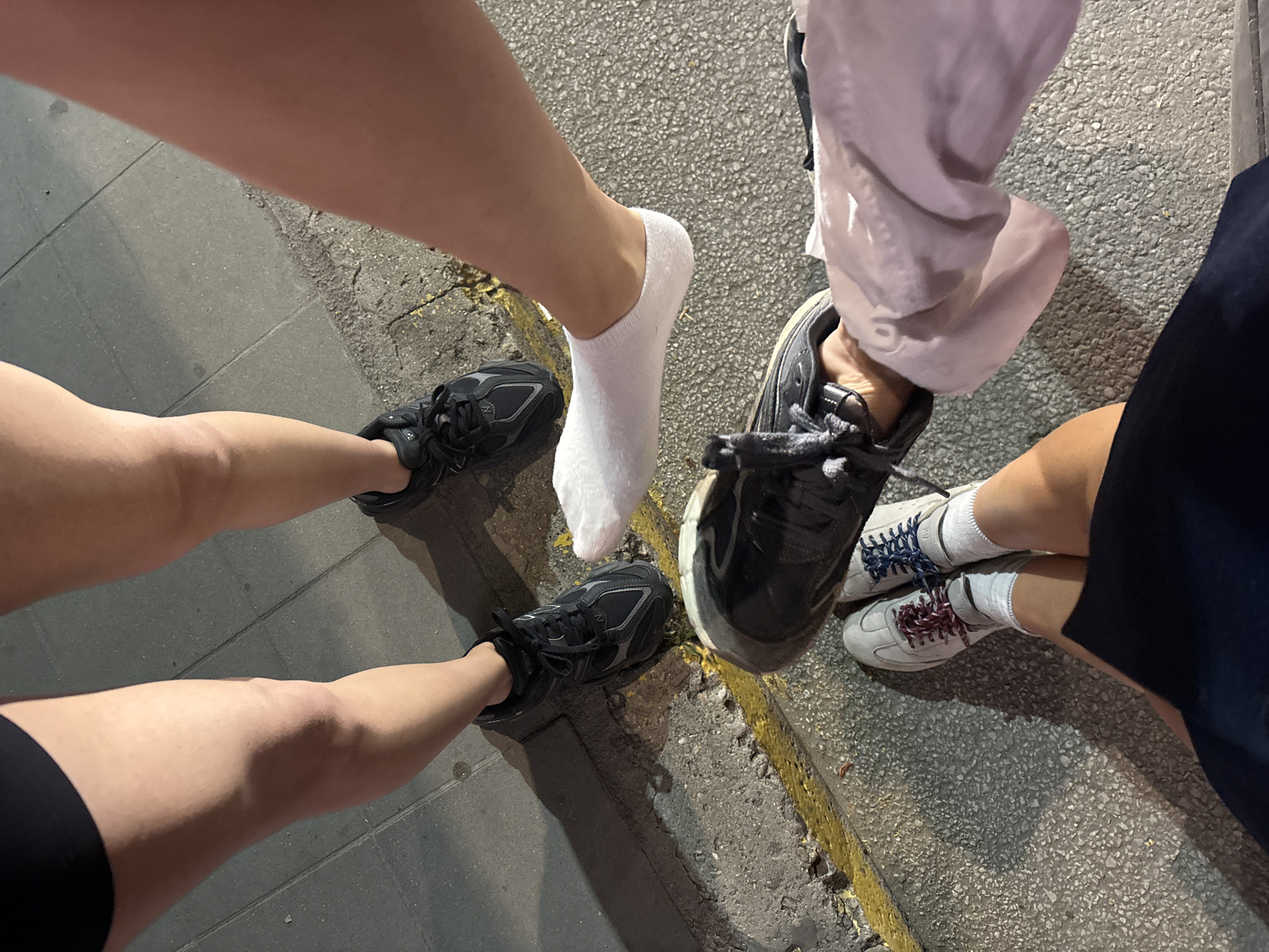 Four people's legs and feet are visible from a top-down perspective, standing on a textured urban surface composed of grey concrete and asphalt, with a distinctive yellow painted line along a raised curb. The scene appears to be a street in Asenovgrad, Bulgaria.
From left to right, the first person's legs are clad in black athletic shorts. They are wearing black athletic shoes with grey accents and white laces.
The second person's legs are bare and appear to be positioned behind the first. They are wearing black athletic shoes with silver accents and grey laces.
The third person is wearing white ankle socks and black athletic shoes with red and blue laces, partially obscured by another person's leg.
The fourth person's legs are partially visible, wearing a light pink long-sleeved top and dark pants. One foot is wearing a white ankle sock and is raised, with the sole of a black shoe clearly visible, adorned with white laces. The other foot is obscured.
The lighting suggests it is evening or nighttime, with artificial light casting shadows. The ground shows signs of wear and tear, typical of a public walkway or street.