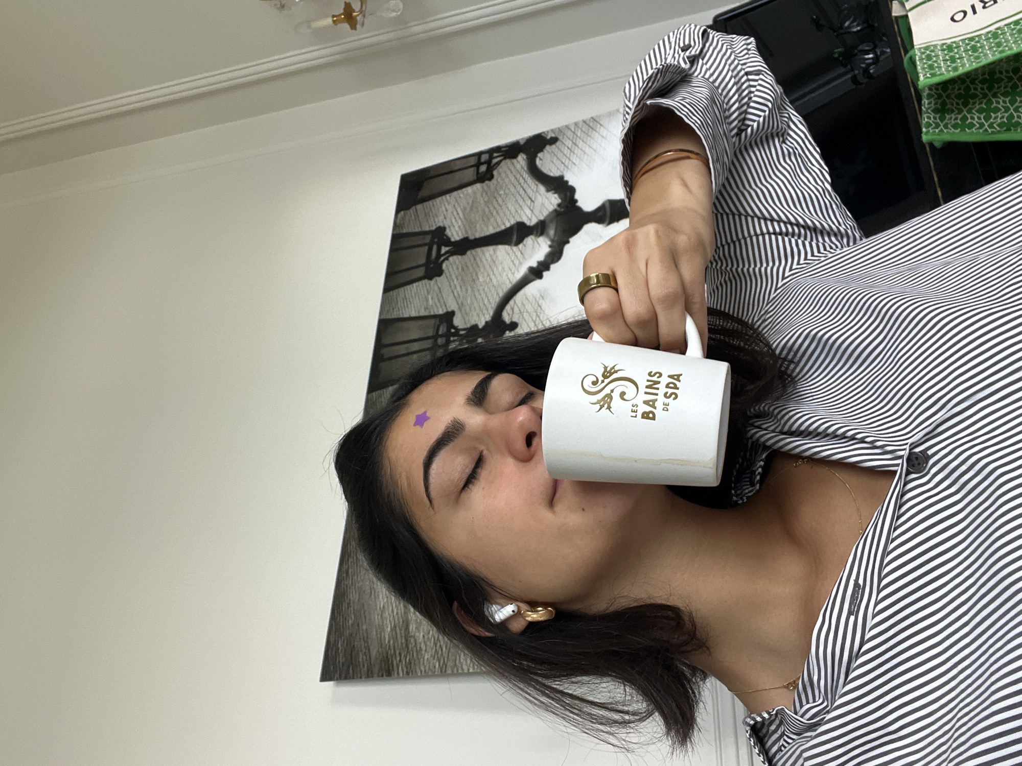 The image, rotated 90 degrees clockwise, presents a close-up shot of a young woman in an indoor setting. She has dark hair, and her eyes are closed in a serene expression, suggesting a moment of relaxation or contemplation. A small purple star sticker is affixed to her forehead. She is holding a white mug with gold lettering to her lips, appearing to drink from it. The mug clearly displays the text "LES BAINS DE SPA" above a decorative gold logo.
The woman is dressed in a black and white striped button-up shirt. Her right hand, holding the mug, features a gold ring on her ring finger and two slender gold bracelets on her wrist. She wears a gold hoop earring and a white earbud in her left ear, and a delicate gold necklace around her neck.
Behind her, a large framed artwork or poster with an intricate black and white pattern, resembling an old lamppost or a decorative metal structure, is mounted on a plain white wall. In the upper right background, a black object, possibly a piece of furniture or a screen, is visible, alongside green and white patterned fabric that shows the word "BIO". A classic-style light fixture with gold details is attached to the ceiling. The scene evokes a sense of peace and personal comfort, potentially in a Parisian home or spa setting.