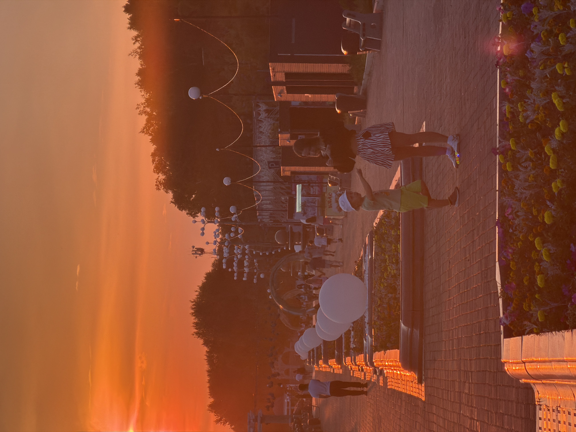 This is a photograph taken during sunset in Birobidzhan, Russia. The sky is a vibrant orange, casting a warm glow over the scene. In the foreground, a brick pathway is lined with planters filled with flowers. Several large, white spherical objects are placed along the path, possibly decorative elements.
Further into the background, a group of people are walking along a promenade. Two children are the central focus in the mid-ground: a young boy in a light-colored t-shirt and green shorts, wearing a white bucket hat, points towards something off-camera. Beside him, a teenage girl in a black top and a zebra-striped skirt is looking down.
Beyond them, there are various structures, including what appear to be small kiosks or stalls. Decorative lights with white spheres are strung along wires above the pathway, adding to the festive atmosphere. Trees line the edge of the park or promenade, silhouetted against the sunset. The overall impression is a lively public space enjoying the last moments of daylight.