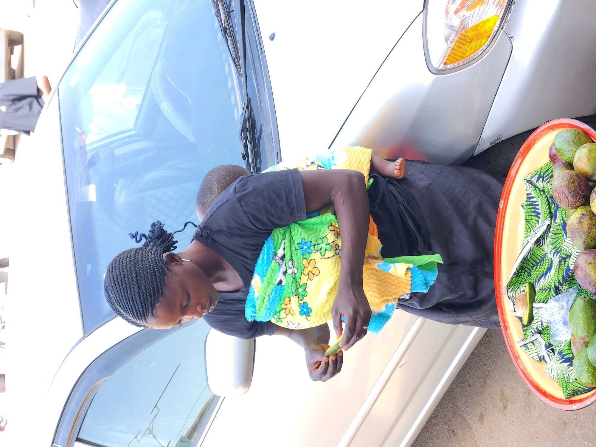 The media file displays a woman carrying a child on her back, standing next to a silver car with a tray of fruits. The scene appears to be outdoors in bright daylight, consistent with a street or market setting in Jos, Nigeria.
The central figure is a woman with her hair styled in small, dark braids, wearing a dark short-sleeved top and a long dark skirt. She carries a baby or toddler on her back, secured with a vibrant yellow, green, and blue patterned blanket adorned with flowers and other motifs. The child's foot is visible. The woman is holding a small, cut piece of fruit, possibly preparing it. Her expression is focused and neutral.
To her right, a round, orange-rimmed tray filled with fruits rests on or near the front passenger side of a silver car. The tray contains numerous avocados, some green and unripe, others with purplish tones indicating ripeness. One avocado is cut open, revealing its pit. The fruits are placed on a green and white patterned fabric, possibly a traditional African print, which lines the tray. The word "GUARANTEEED" is partially visible on this fabric.
The silver car serves as a backdrop, with its front headlight and bumper visible on the right. The car's windshield reflects the bright, clear sky, indicating sunny weather. The overall impression is that of a street vendor or market stall where the woman is selling fruits while attending to her child.
