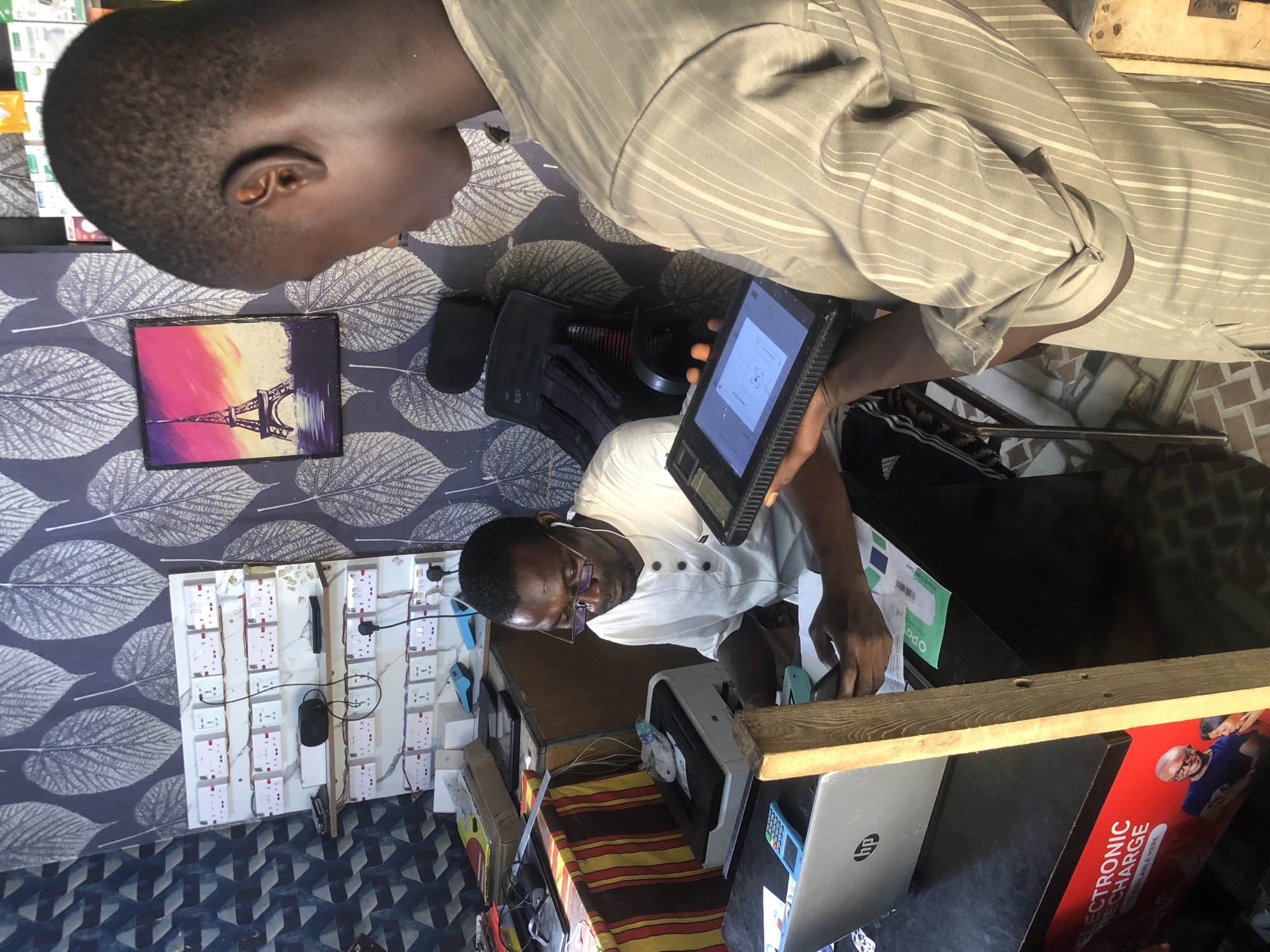 This image captures a scene inside a small shop in Ado, Nigeria, likely a telecommunication or electronics store. Two young men are engaged in a transaction. The man on the left, wearing a white t-shirt and glasses, is seated at a table, looking down at his mobile phone. In front of him is a silver laptop with an HP logo and a light blue calculator. A printer is also visible on the table, along with some papers. He appears to be focused on his phone and possibly inputting information.
The man on the right, wearing a striped, light-colored shirt, stands and holds a black tablet device. He is looking down at the tablet, likely displaying information to the man seated or about to show him something on the device. His right hand is holding the tablet, and his left hand is partially visible, also near the device.
The background reveals the shop's interior. The wall is decorated with a dark wallpaper featuring a repeating pattern of light gray leaves. To the left, a white board is mounted with numerous electrical outlets and sockets, suggesting an electrical supply aspect to the business. A small framed picture of the Eiffel Tower hangs on the wall behind the man on the right. The overall lighting suggests an indoor environment, possibly with overhead lights illuminating the scene. The flooring appears to be tiled. The atmosphere suggests a routine business interaction within a localized urban setting in Nigeria.