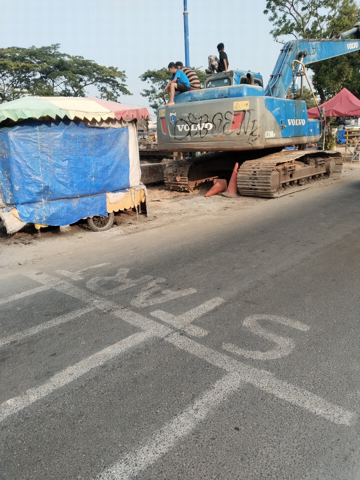 In West Jakarta, Indonesia, a blue Volvo excavator sits idle on a dirt road beside a temporary stall covered in blue tarp and a colorful awning. Two young boys are perched on the excavator's body, one in a blue and striped shirt, the other in a black shirt. White markings, possibly indicating a crosswalk or lane division, are painted on the asphalt in the foreground. Traffic cones are visible near the excavator's tracks. Trees and a clear sky form the background.