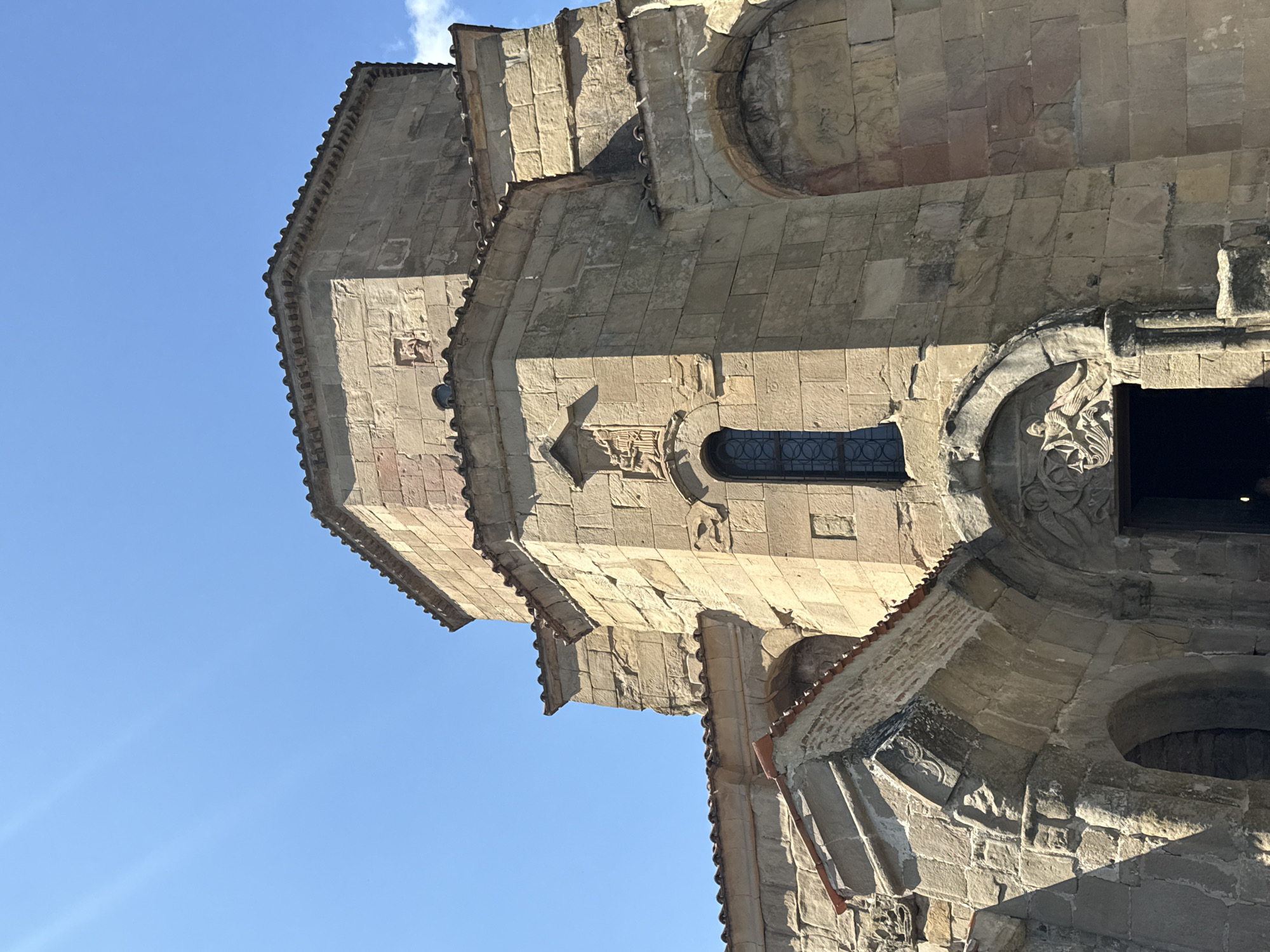 This is a low-angle, eye-level, outdoor, full shot of the Jvari Monastery in Mtskheta, Georgia, under a clear blue sky. The ancient stone structure features a large, multi-faceted dome and intricately carved details above the entrance and windows. The stonework is weathered and shows signs of age, with some areas of erosion and discoloration. The roof is tiled with dark, reddish-brown tiles. Sunlight casts shadows on the building, highlighting the texture of the stone. The entrance is an arched doorway with ornate carvings above it. The overall scene conveys a sense of historical significance and architectural grandeur.