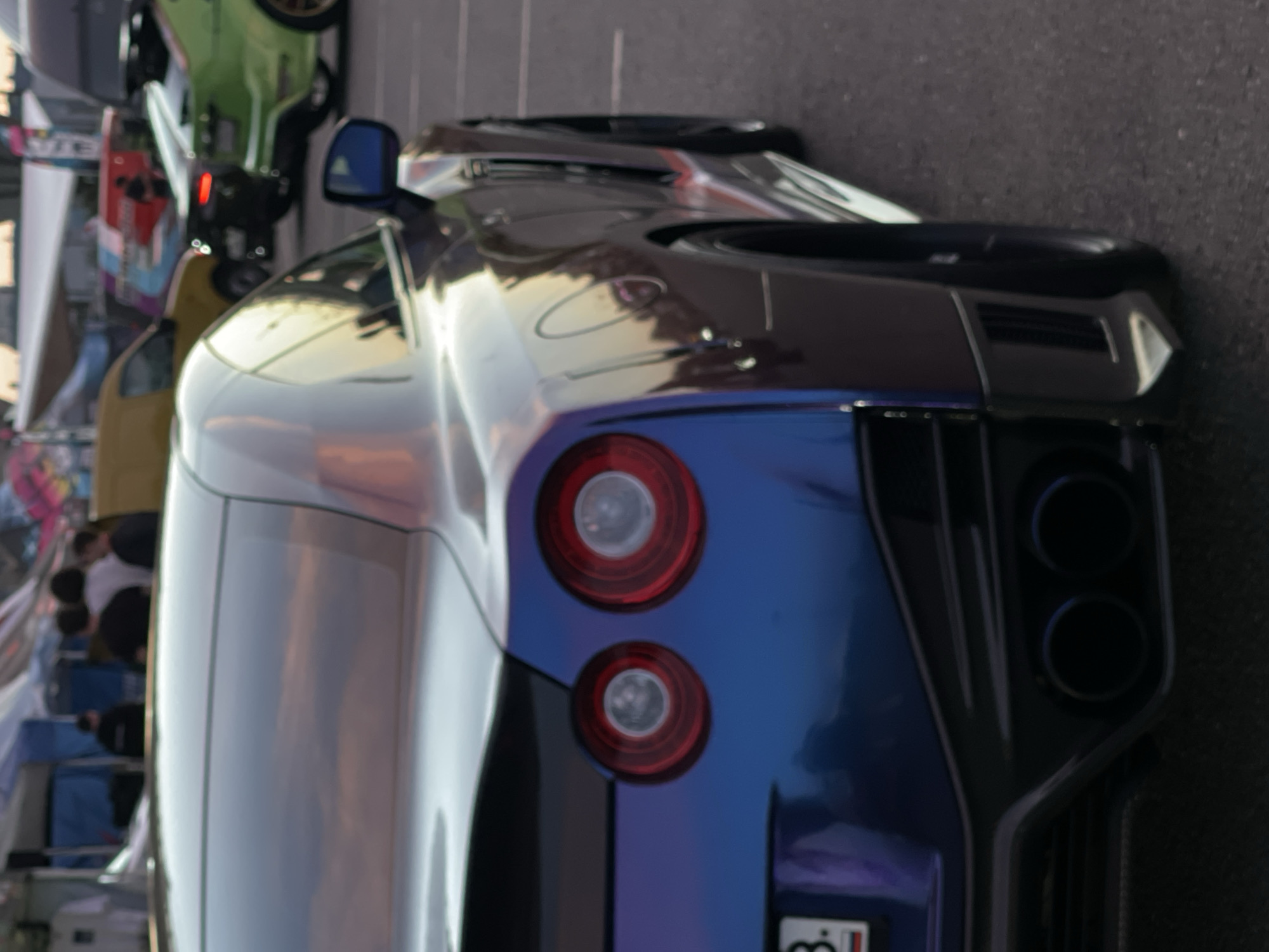 An eye-level, close-up shot shows the rear passenger side of a dark iridescent purple and blue sports car parked on asphalt. The car, possibly a Nissan GT-R, is angled away from the camera, with its tail lights, exhaust pipes, and rear bumper visible. The tail lights are circular and red with white centers. The bumper is black and features a diffuser with two large circular exhaust tips. A portion of the license plate with the number "3." is visible on the left side of the bumper. The car's paint reflects the sky and surrounding environment. In the background, out of focus, are tents, people, and other vehicles, suggesting an outdoor event or car show. The lighting indicates it is either early morning or late afternoon.