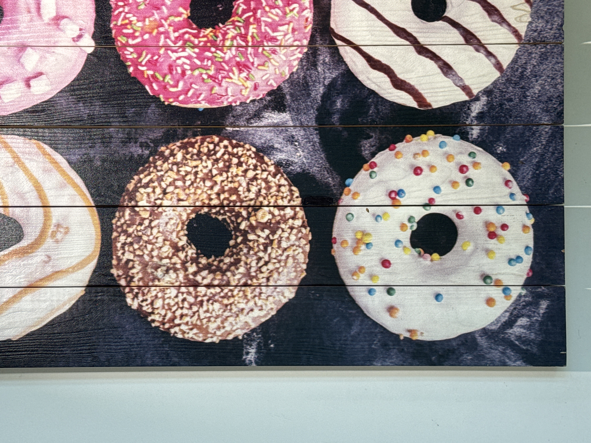 This image is a close-up shot of a piece of wall art featuring six donuts arranged in a 3x2 grid. The donuts are depicted on a dark, textured background that resembles wood grain. The donuts themselves vary in decoration: the top row shows a pink donut with white frosting and sprinkles on the right, and a donut with orange stripes and white frosting on the left. The middle row has a chocolate donut covered in nuts on the left and a pink donut with rainbow sprinkles on the right. The bottom row displays a white donut with colorful sprinkles on the left and a white donut with dark brown stripes on the right. The artwork is mounted on a dark vertical surface, and a light blue wall is visible to the left of the artwork. The overall impression is a colorful and decorative display.