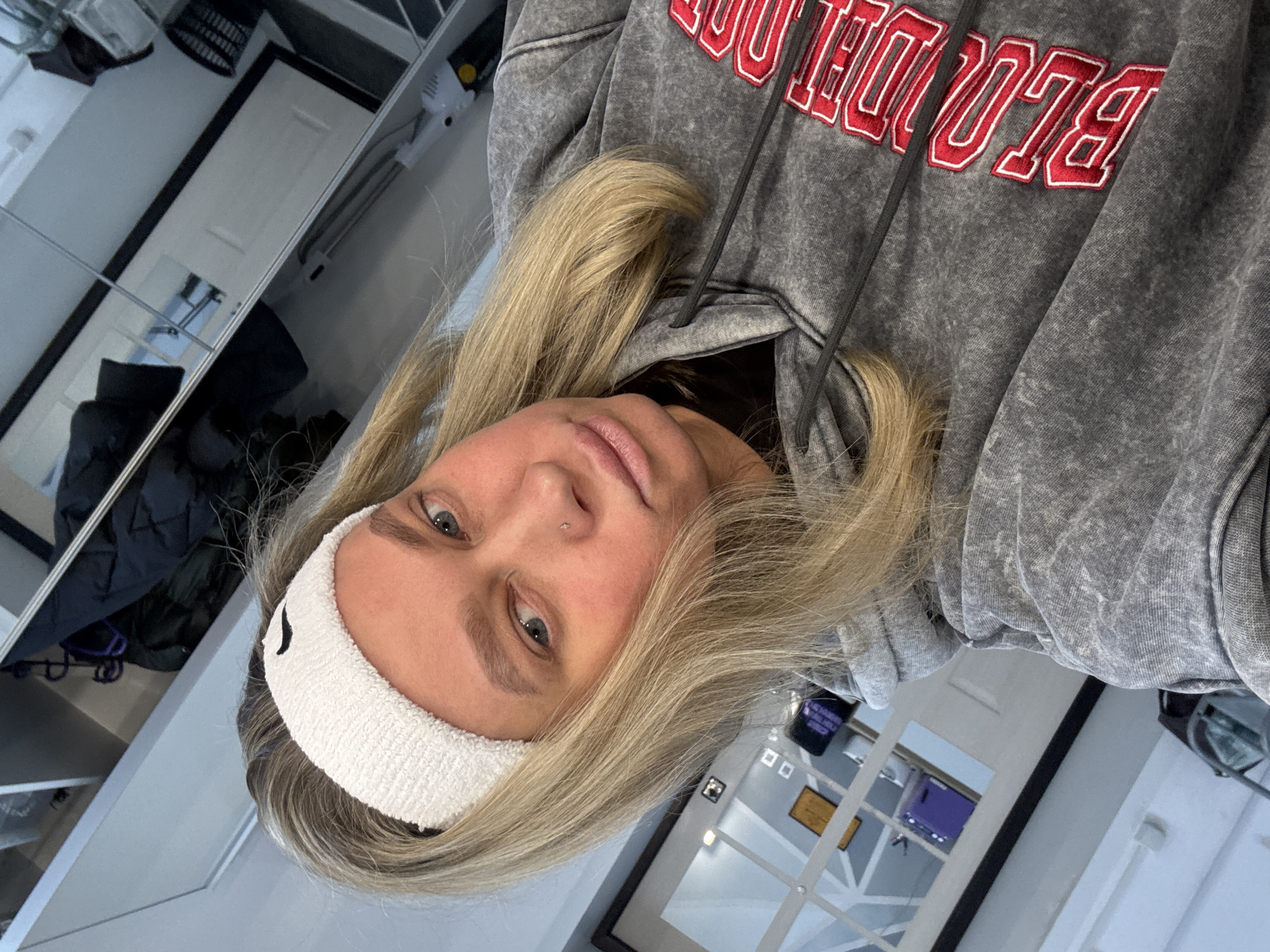 An eye-level selfie shows a young woman with long blonde hair and light-colored eyes, taken indoors. She has a neutral and calm expression, looking directly at the camera. She wears a white terry cloth headband on her forehead and a small nose piercing. Her attire consists of a grey marled hoodie with red and white embroidered text across the chest. The visible text on the hoodie clearly spells "BLOOD" followed by "BLVD", likely an abbreviation for Boulevard.
The background reveals an indoor setting, possibly a gym, changing room, or a modern space with various reflective surfaces, possibly mirrors or glass panels, giving a sense of depth and reflecting parts of the room. Reflections include a white door, dark clothing hanging on a rack or hook, and what appears to be shelves or cubbies. In one reflection, a dark, possibly purple, object or screen is visible. The lighting is even, suggesting an interior during the daytime. The general ambiance is clean and functional. The location is in Perm, Russia.