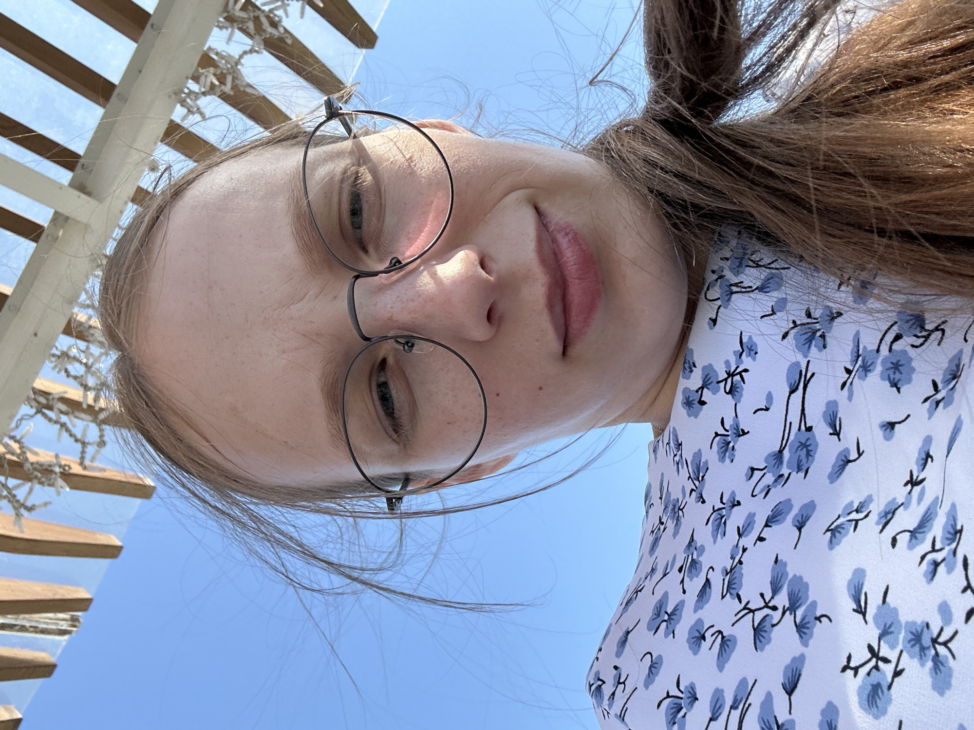 The image shows a close-up, low-angle selfie of a young woman with blonde hair and freckles, wearing glasses and a white dress with a blue floral pattern. She is looking towards the camera with a slight smile. The background consists of a clear blue sky and a wooden structure overhead, possibly part of a canopy or pergola. The lighting suggests it is daytime, likely outdoors. The location is identified as Металлплощадка, Russia.
