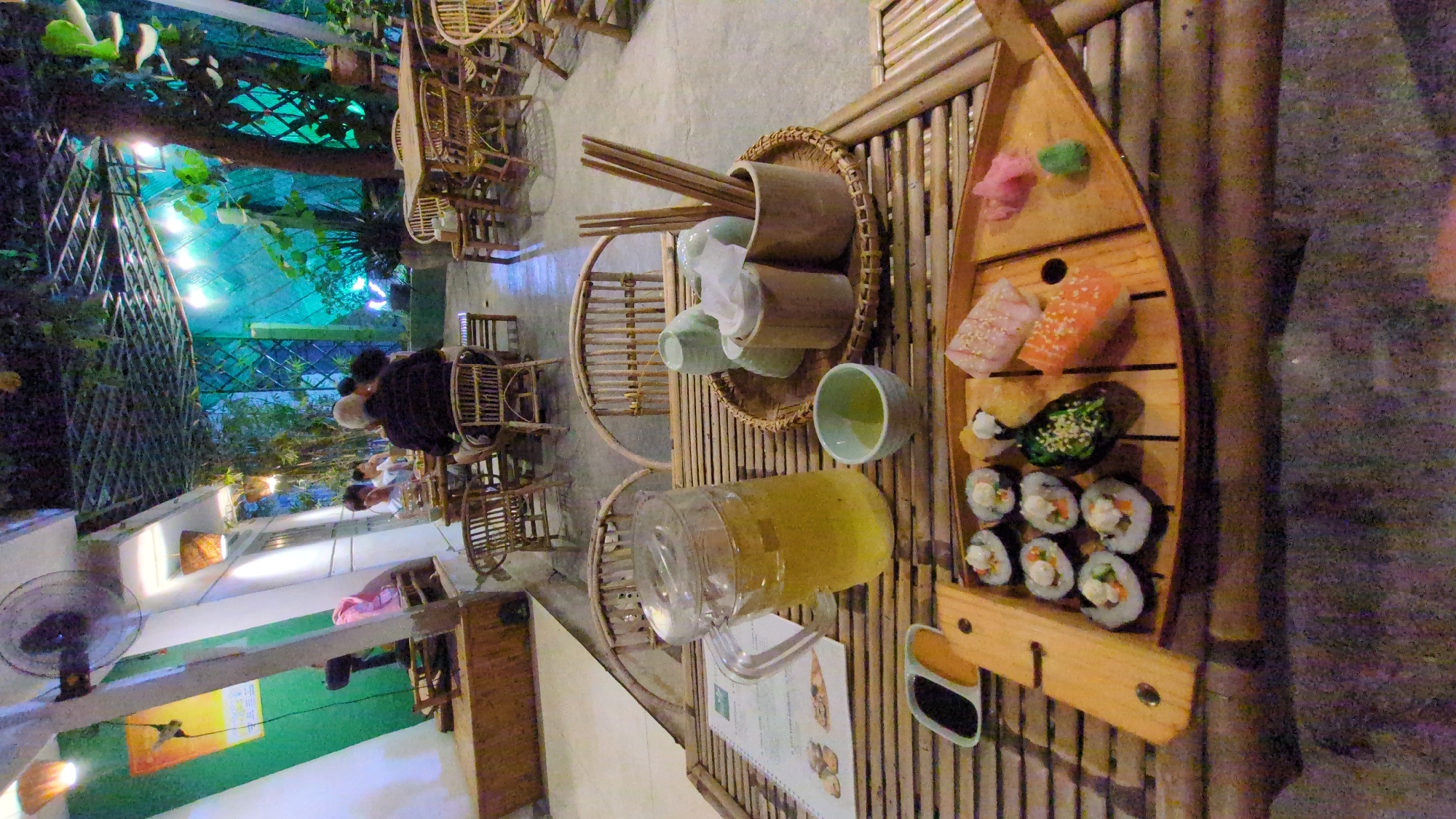 The scene depicts a restaurant setting in Phan Rang – Tháp Chàm, Vietnam. A bamboo table is laden with a wooden boat-shaped tray filled with assorted sushi, including maki rolls and nigiri, accompanied by pickled ginger and wasabi. There's a pitcher of liquid, possibly tea or juice, and a set of tea cups with chopsticks in a bamboo holder. A menu is also visible on the table. In the background, people are seated at other bamboo tables and chairs, with a view of the outdoor setting including a fence, foliage, and overhead lighting. A standing fan is visible in the lower left corner.