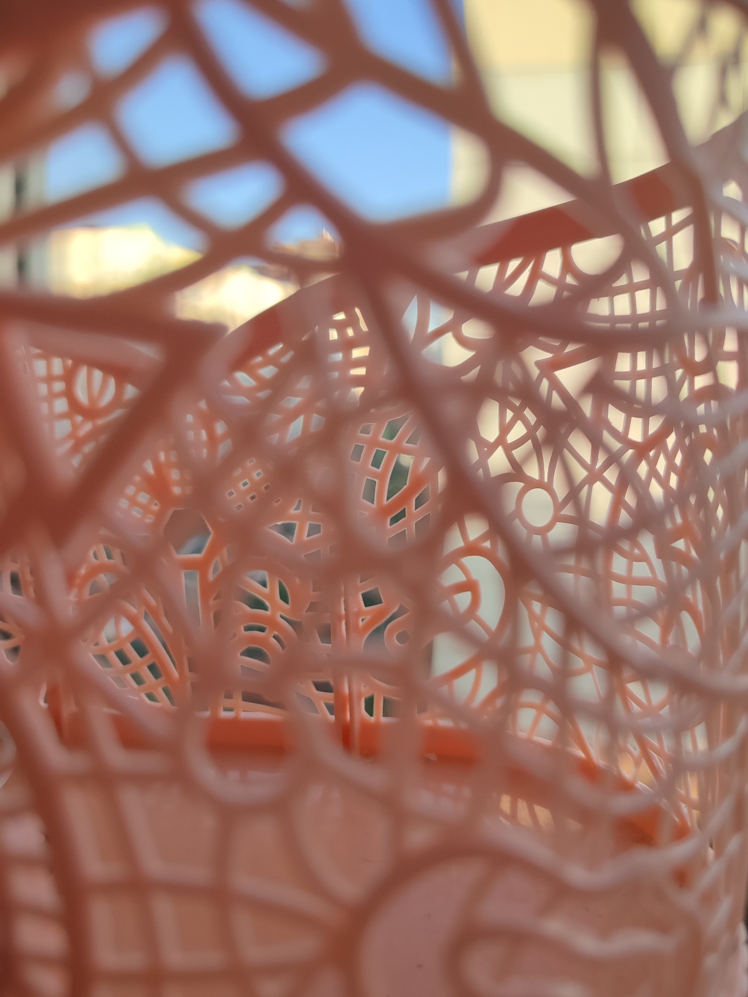 The image shows a close-up, out-of-focus view of a pink plastic basket. The basket has an intricate woven pattern with circular and hexagonal openings. Through the openings, a blurred background is visible, consisting of a light blue sky, pale yellow buildings, and some green foliage. There are no people or any other identifiable objects in the scene. The image's focus is entirely on the textured surface of the basket and the abstract patterns it creates. There is no information in the image that would indicate its location in the city of Voronezh, Russia.