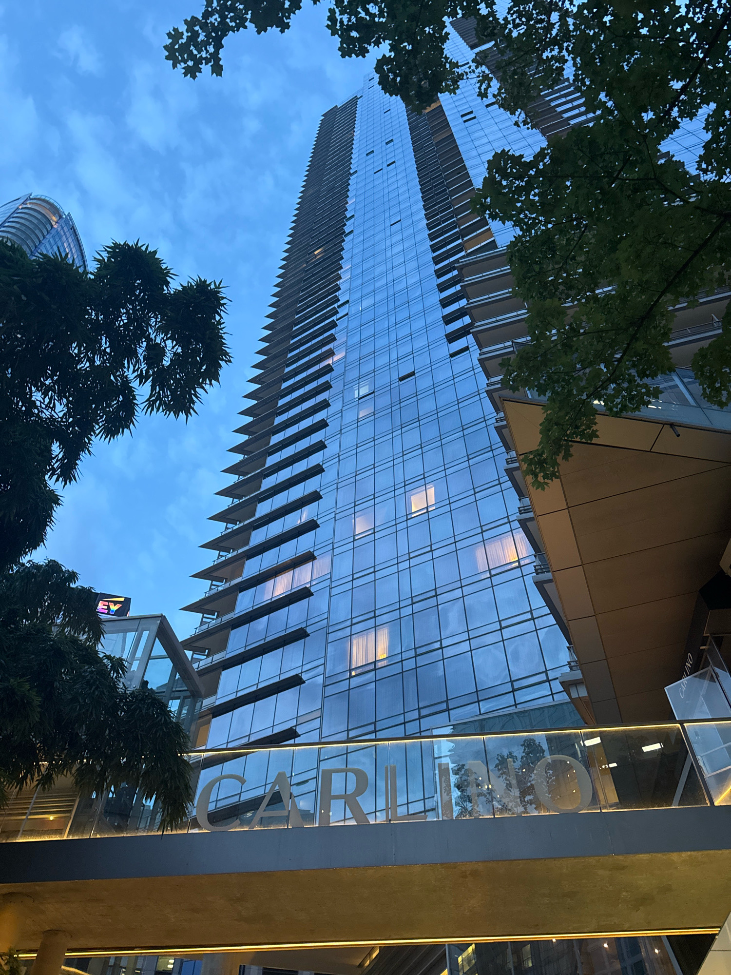 The image shows a modern glass-covered skyscraper in Vancouver, Canada, possibly at dusk. The building's facade reflects the cloudy blue sky and the surrounding greenery of trees. In the foreground, a walkway with the word "CARLINO" spelled out in large letters on its railing is visible. Another building with a curved facade can be seen in the background to the left. The overall scene suggests an urban setting with contemporary architecture.