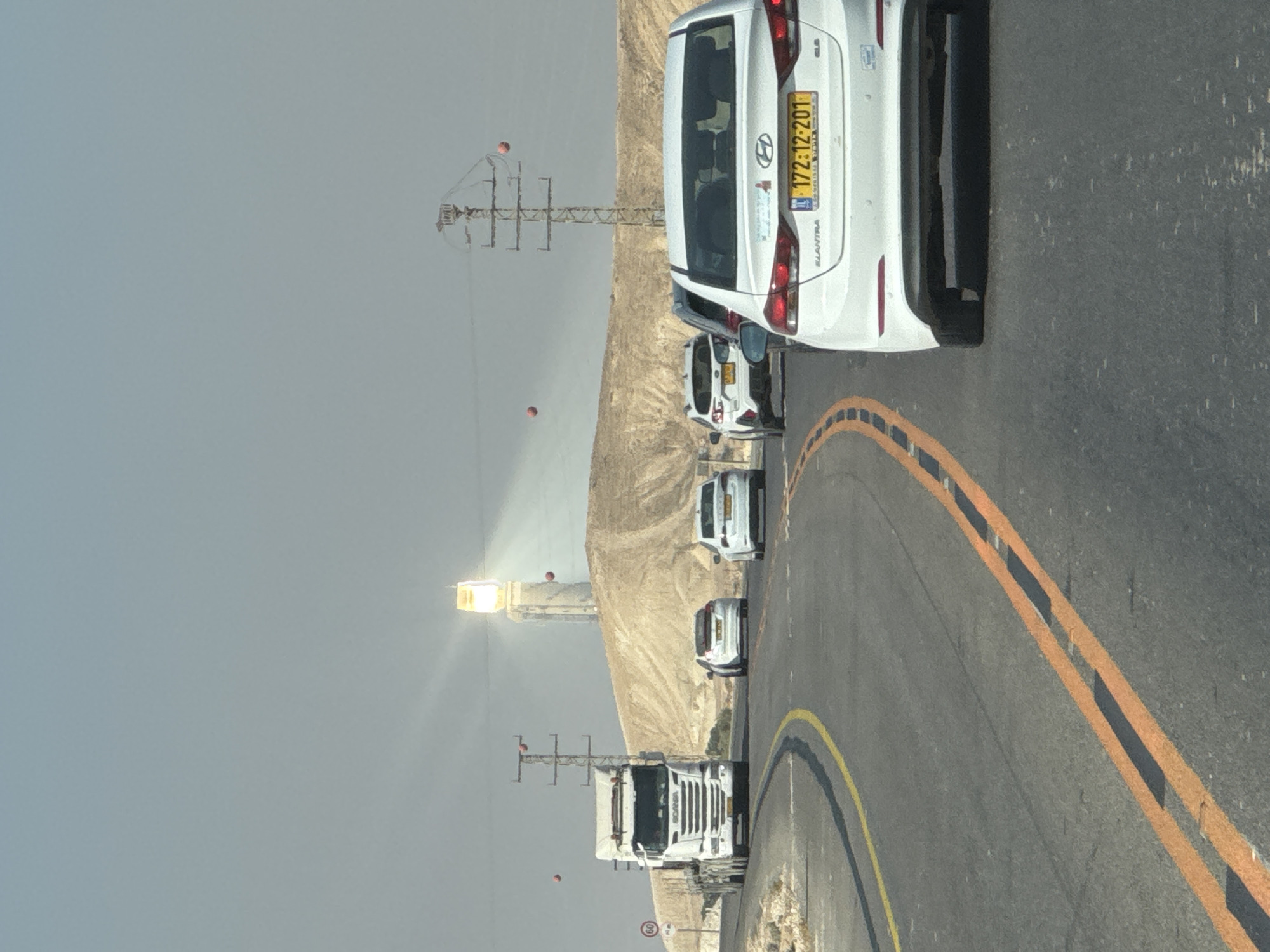 The image shows a highway scene in Ramat Negev Regional Council, Israel, with a white Scania truck on the left and several cars on the right, including a white Hyundai Elantra in the foreground. The vehicles are traveling on a road with clear lane markings. In the background, a tall tower with a bright light at the top is visible against a hazy sky. There are also power lines with red spherical markers and a sign indicating a speed limit of 60. The landscape appears arid, with hills and sparse vegetation. The overall impression is of a journey through a desert region.