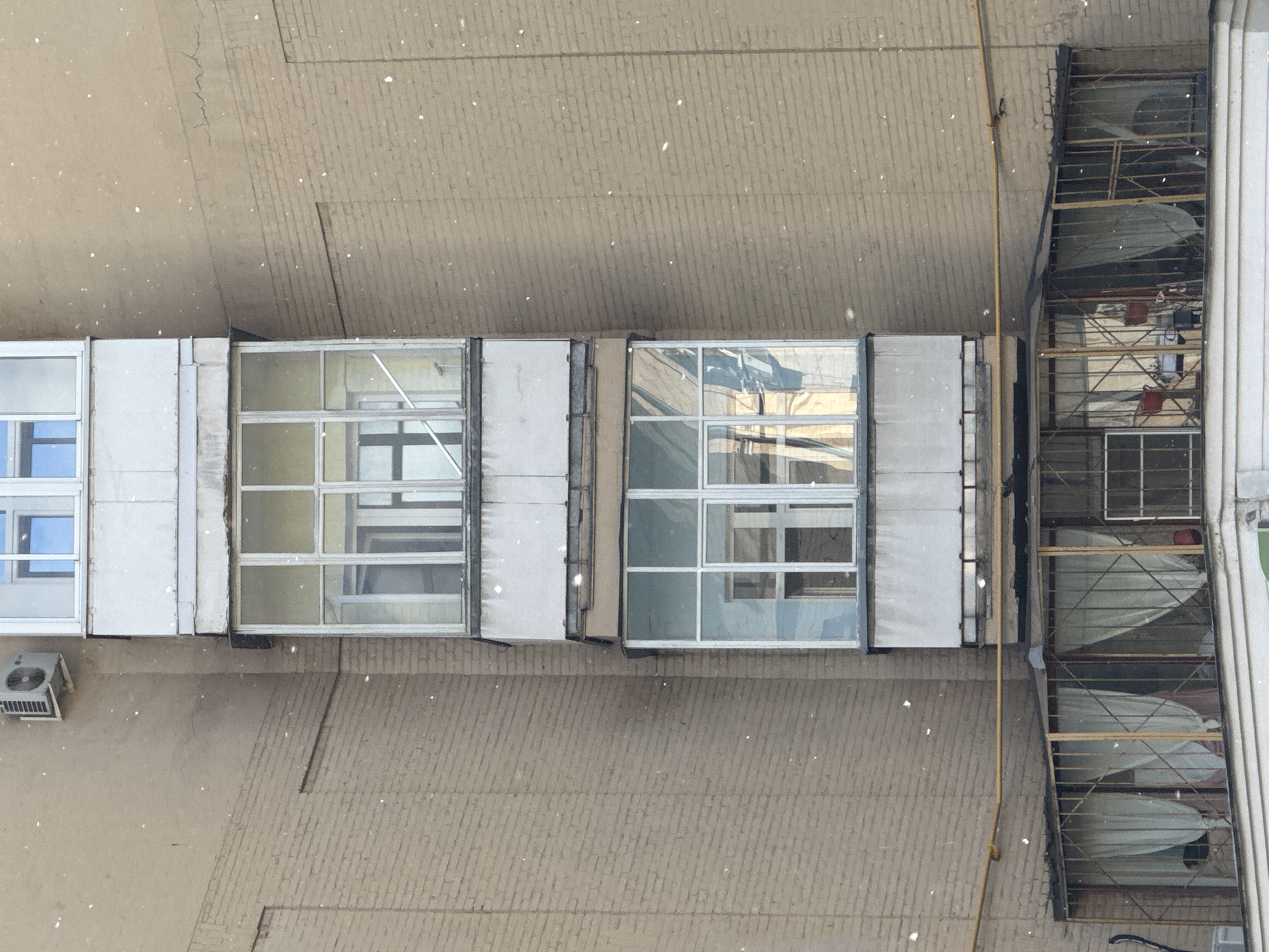 This high-angle shot, taken from above, provides a view of an urban building exterior or courtyard in Moscow, Russia, during daylight hours.
The central focus is on two prominent structures. On the left, a light-colored, elongated enclosure, possibly a walkway or building extension, features multiple rectangular windows with dark frames. These windows reflect various elements of the surroundings. Below this structure, on the textured ground, a grey outdoor air conditioning unit is mounted on the far left.
Adjacent to this, on the right, is a multi-tiered metal framework, resembling a balcony or utility scaffolding. Several sections of this structure are covered with translucent white material, possibly fabric or plastic sheets, which partially obscure the interior. Faintly visible within this framework are two small red buckets. A distinct yellow pipe runs vertically along the left edge of this framework and extends downwards.
The ground or wall surface beneath and around these structures is a uniform light brown or beige, exhibiting a subtle, patterned texture that suggests brickwork or concrete. Small, bright white specks are scattered across the entire image, particularly noticeable against the ground, indicating light snowfall or airborne particles. The lighting is bright and even, consistent with daytime. No individuals or readable text are visible in the scene.
