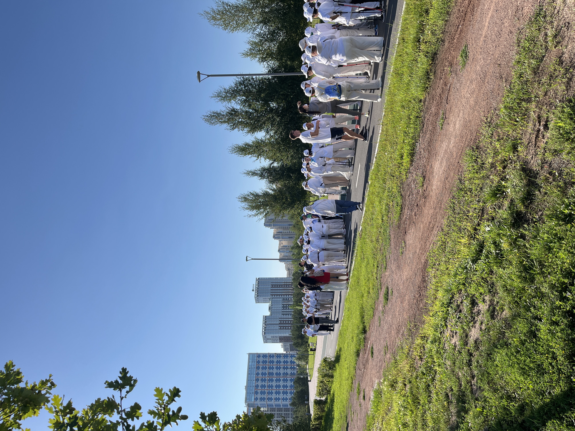 In Astana, Kazakhstan, a large group of people, predominantly dressed in white outfits and hats, are gathered on a paved path in a park-like setting. Many are holding walking poles, suggesting they are participating in a group walking activity, possibly Nordic walking. The group is arranged in several rows, facing generally towards the right side of the frame. Some individuals in the front are engaged in conversation, with one person speaking into a microphone. In the background, modern high-rise buildings are visible, indicating an urban environment. Tall, slender trees line the path behind the group, and lush green grass covers the ground. The sky is clear and blue, suggesting a sunny day. The overall scene depicts an organized outdoor recreational event in a contemporary urban park.
