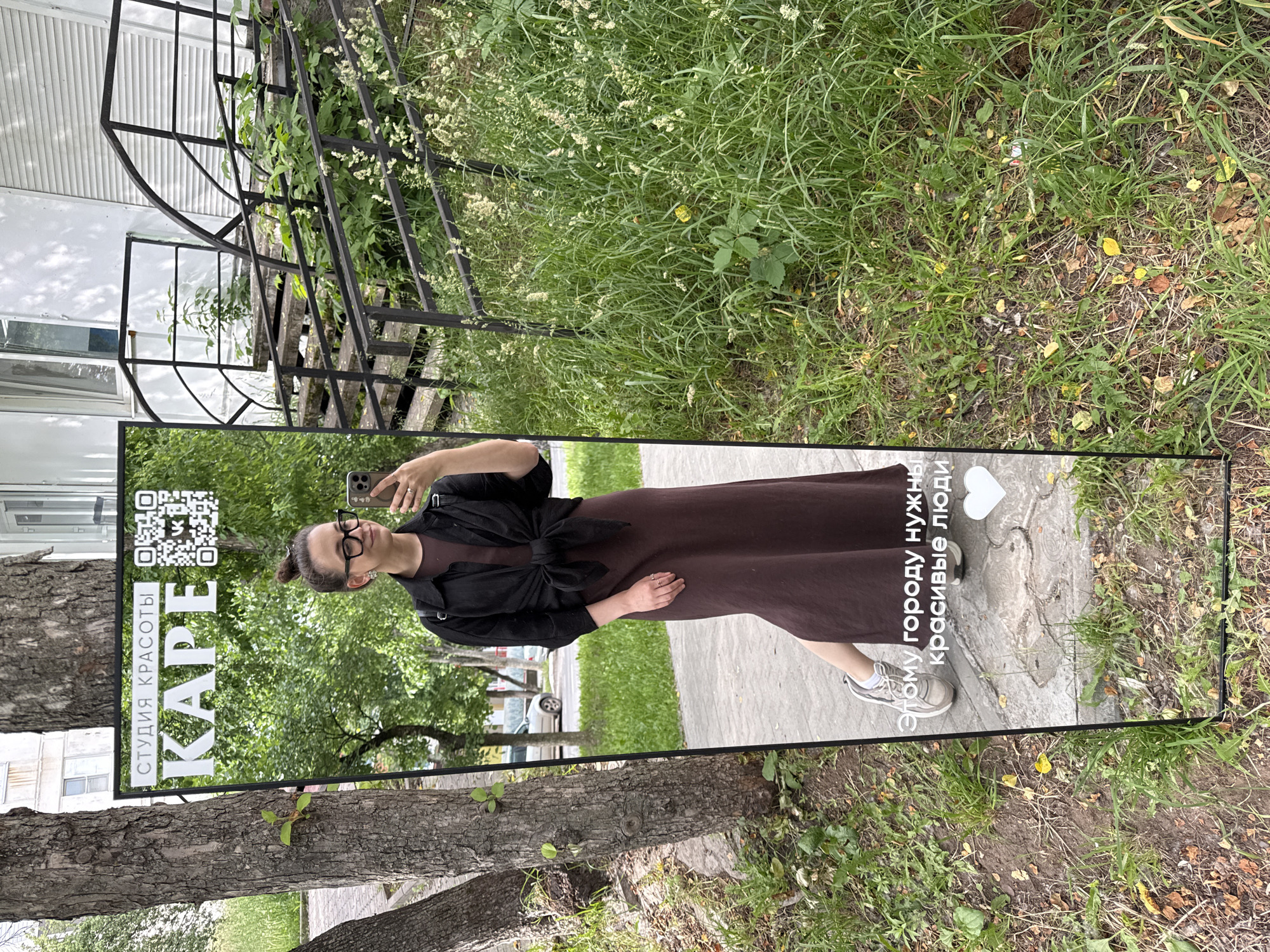A full-length mirror stands outdoors, propped up in a grassy area next to a tree. The mirror reflects a young woman wearing glasses, a black jacket tied around her waist over a long brown dress, and white sneakers. She is holding a smartphone in her right hand, appearing to take a selfie. Text is visible on the mirror, with the top portion reading "СТУДИЯ КРАСОТЫ КАРЕ" (Beauty Studio KARE) and a QR code. Below this, in Russian, is the phrase "Этому городу нужны красивые люди" (This city needs beautiful people), accompanied by a white heart symbol. The background in the reflection shows a building with stairs and lush green vegetation. The scene is set in Chaikovsky, Russia, specifically on a sidewalk area with trees and grass.