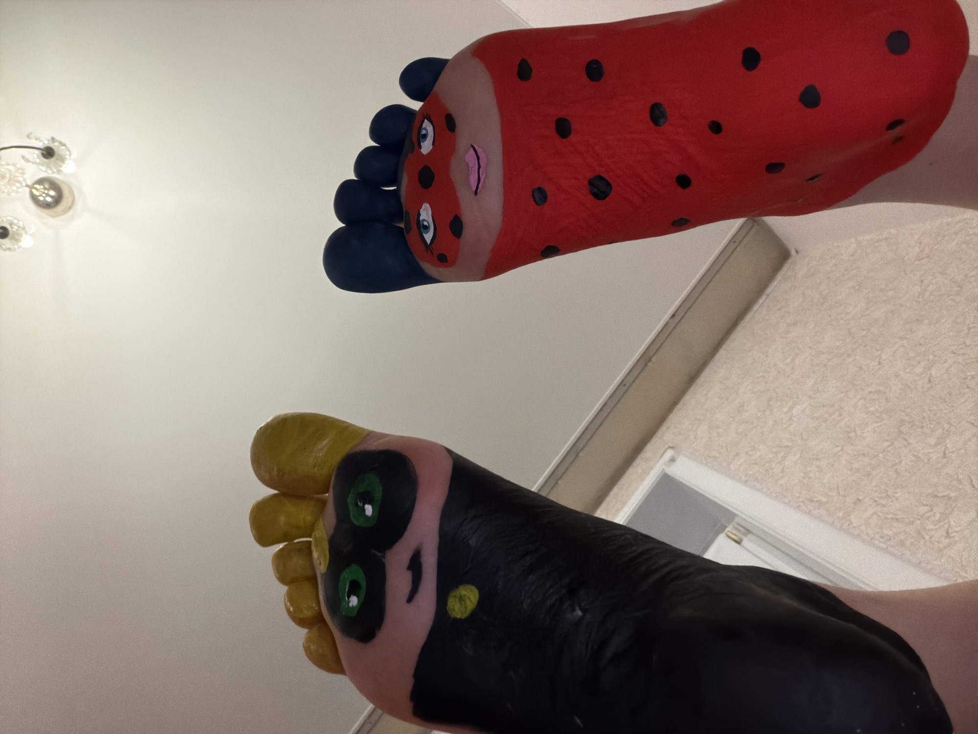 The image shows two feet, each painted to resemble characters from the animated series "Miraculous: Tales of Ladybug & Cat Noir." The right foot is painted red with black spots to represent Ladybug, and the toes are painted blue. The left foot is painted mostly black, with the toes painted yellow and two green eyes painted on the upper part of the foot to represent Cat Noir. The feet are angled upwards towards the camera against a white ceiling and wall. A light fixture is visible in the background. There is no information available to establish the location to be Perm, Russia.