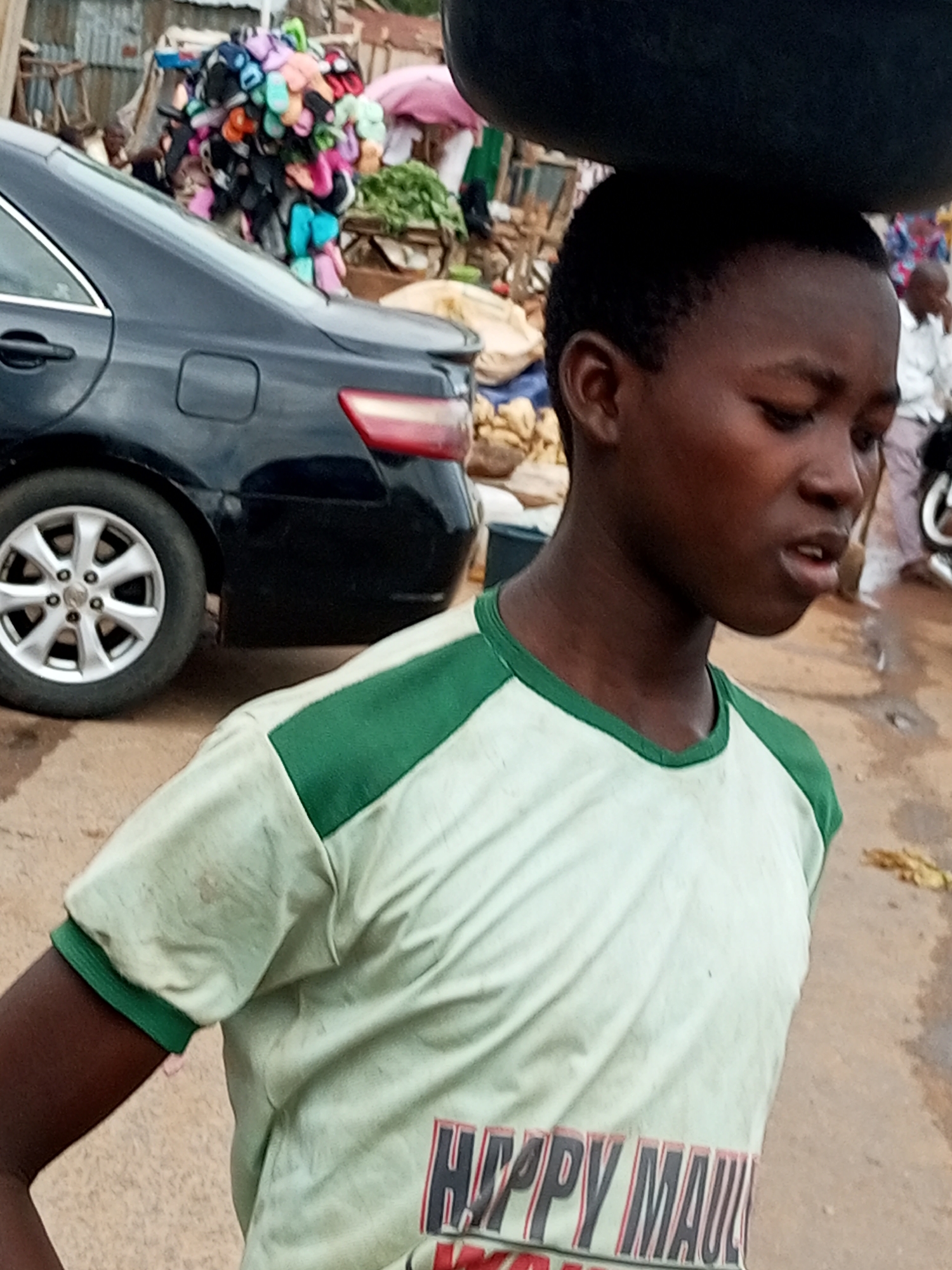 A young person is shown from the chest up, facing to the right and looking down. They are carrying a large, dark, circular object on their head. They wear a light-colored t-shirt with green trim around the collar and sleeves. The t-shirt has a graphic design on the lower part, reading "HAPPY MAUL" with red and black lettering. The background suggests an outdoor market setting in Tempure, Nigeria, with a dark sedan parked behind the person and a stall piled with colorful footwear to the left. Other market elements, including people and produce, are visible in the background. The ground appears to be dirt or a similar unpaved surface.