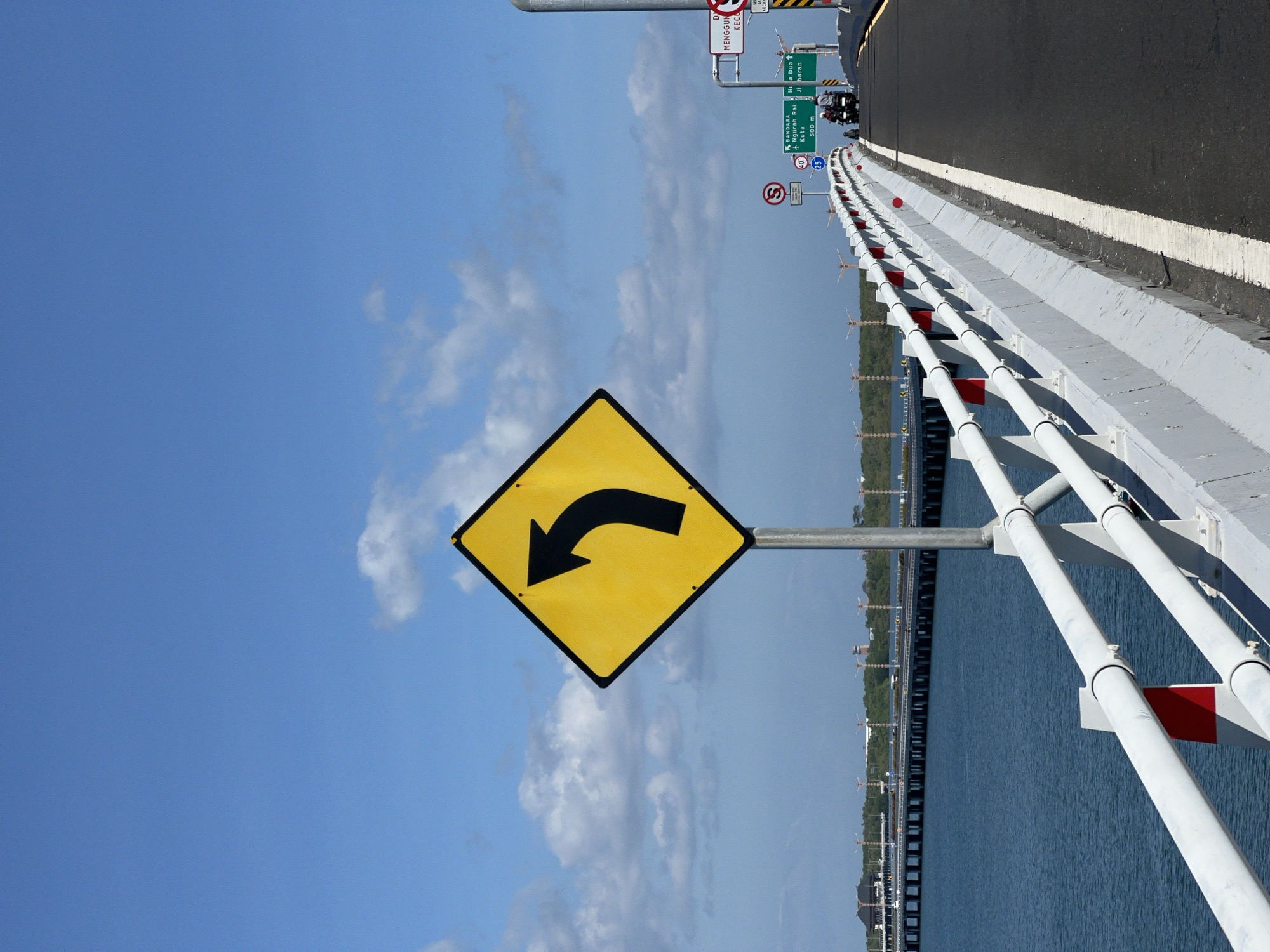The image depicts a portion of a bridge in Kuta, Indonesia. A yellow, diamond-shaped traffic sign with a black curved arrow pointing left is prominent on the left side. The bridge has a grey concrete barrier with a white stripe and a white metal railing. The bridge spans a body of water, with land visible in the background. Above, a blue sky with scattered clouds fills the frame. On the right edge of the bridge, there are road signs, some motorcycles and speed limit signs.