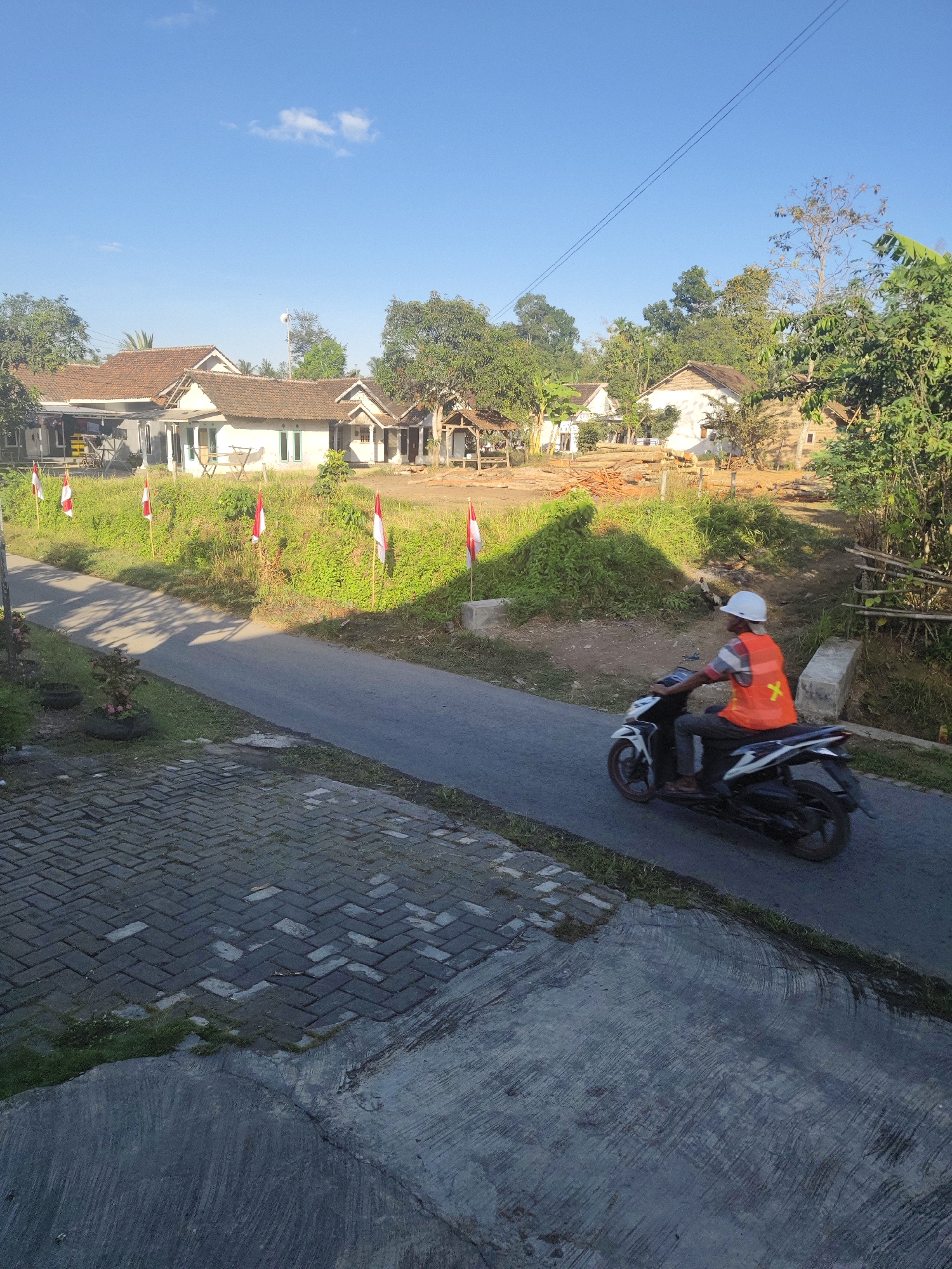 A motorcyclist wearing a white hard hat and an orange safety vest is riding a white motorcycle on a paved road. The road is lined with green grass and several red and white Indonesian flags on poles. In the background, there are several houses with tiled roofs, surrounded by trees and vegetation. The sky is blue with a few white clouds. The scene appears to be a rural or suburban area in Bangsalsari, Indonesia, possibly during a festive occasion given the flags.